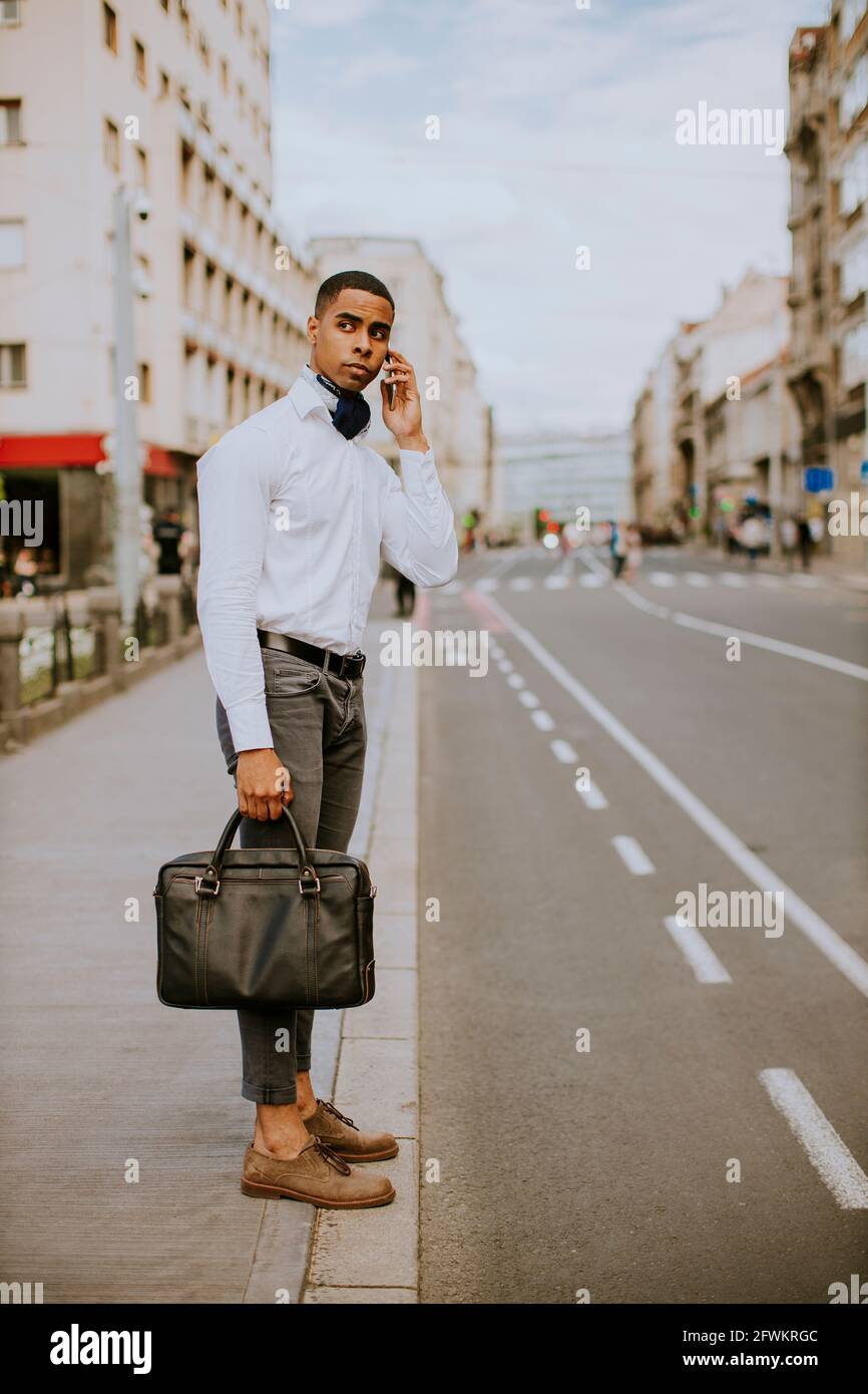 Bel giovane uomo d'affari afro-americano che usa un telefono cellulare mentre attesa di un taxi in attesa di un taxi su una strada Foto Stock