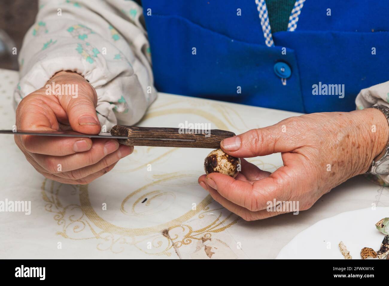Le mani di una signora anziana irriconoscibile che batte un bollito quail uovo con il manico di un coltello per rimuovere la conchiglia per preparare un pasto Foto Stock