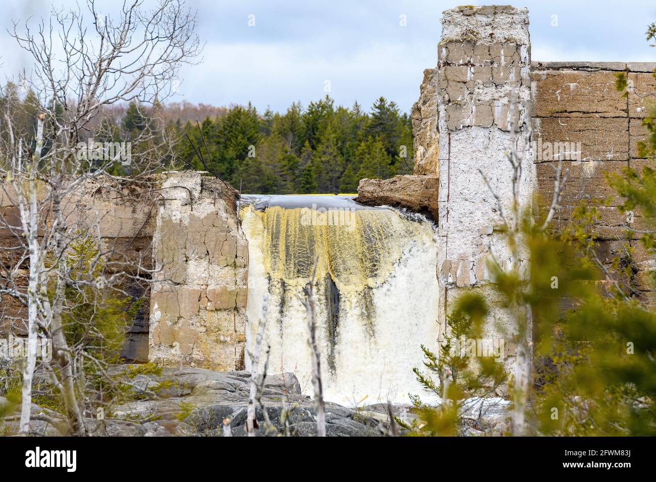 L'acqua scorre su una vecchia diga rotta. La diga è sbriciolare, ed è vista attraverso gli alberi. Cielo sovrastato. Foto Stock