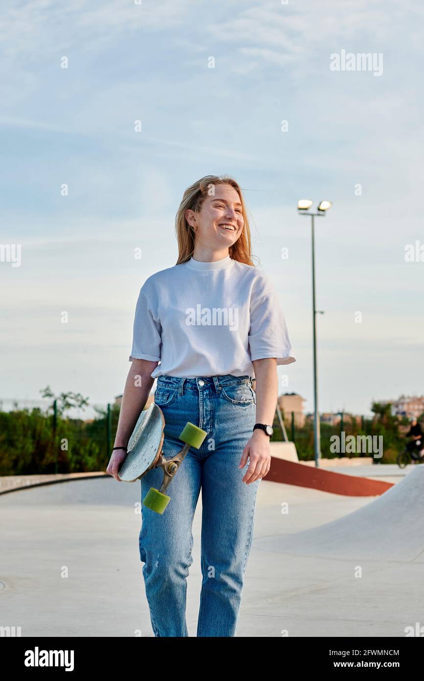 Giovane donna bionda sorridente e divertente tenere uno skateboard Foto Stock