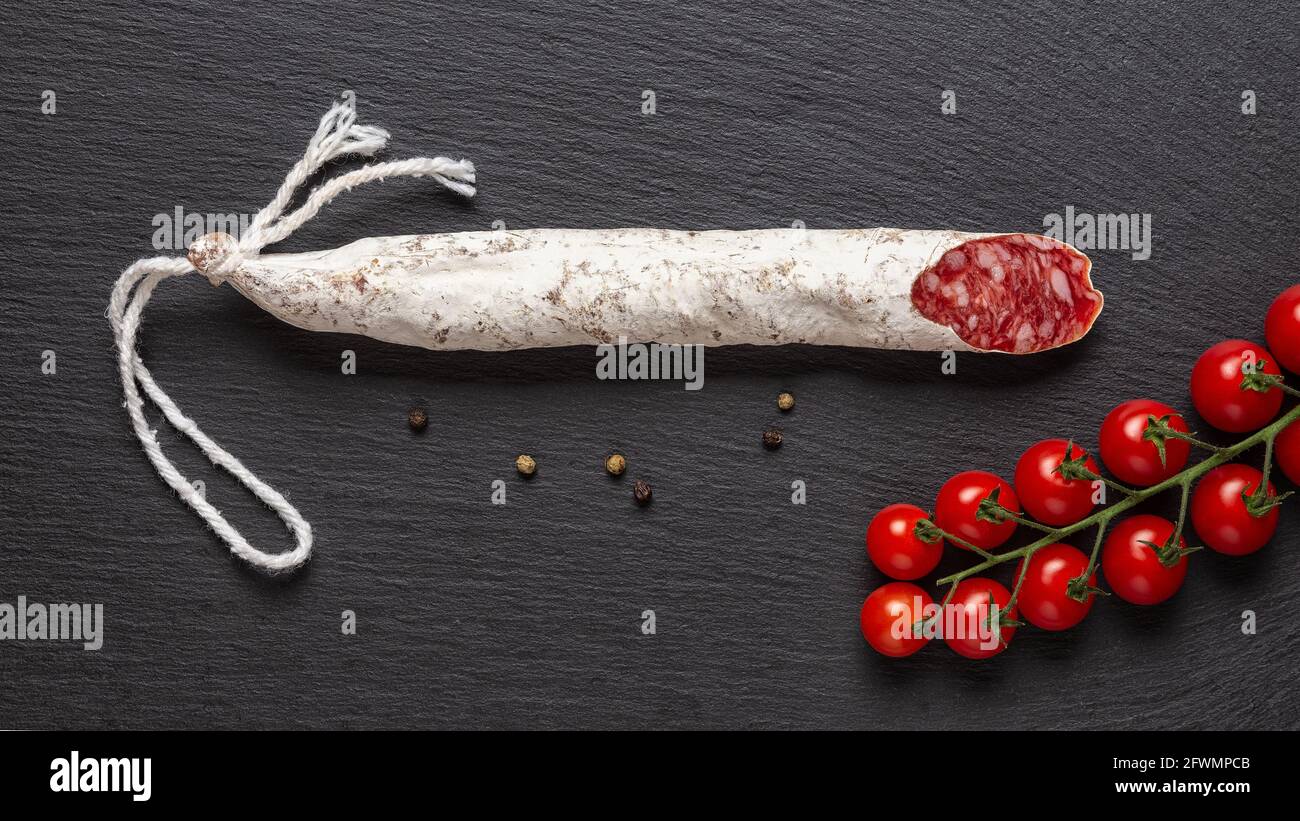 Sottile salsiccia di fueta catalana e mazzo di pomodori ciliegia rossa su una superficie di ardesia nera. Ampia immagine del tradizionale salame spagnolo di maiale stagionato a secco. Foto Stock