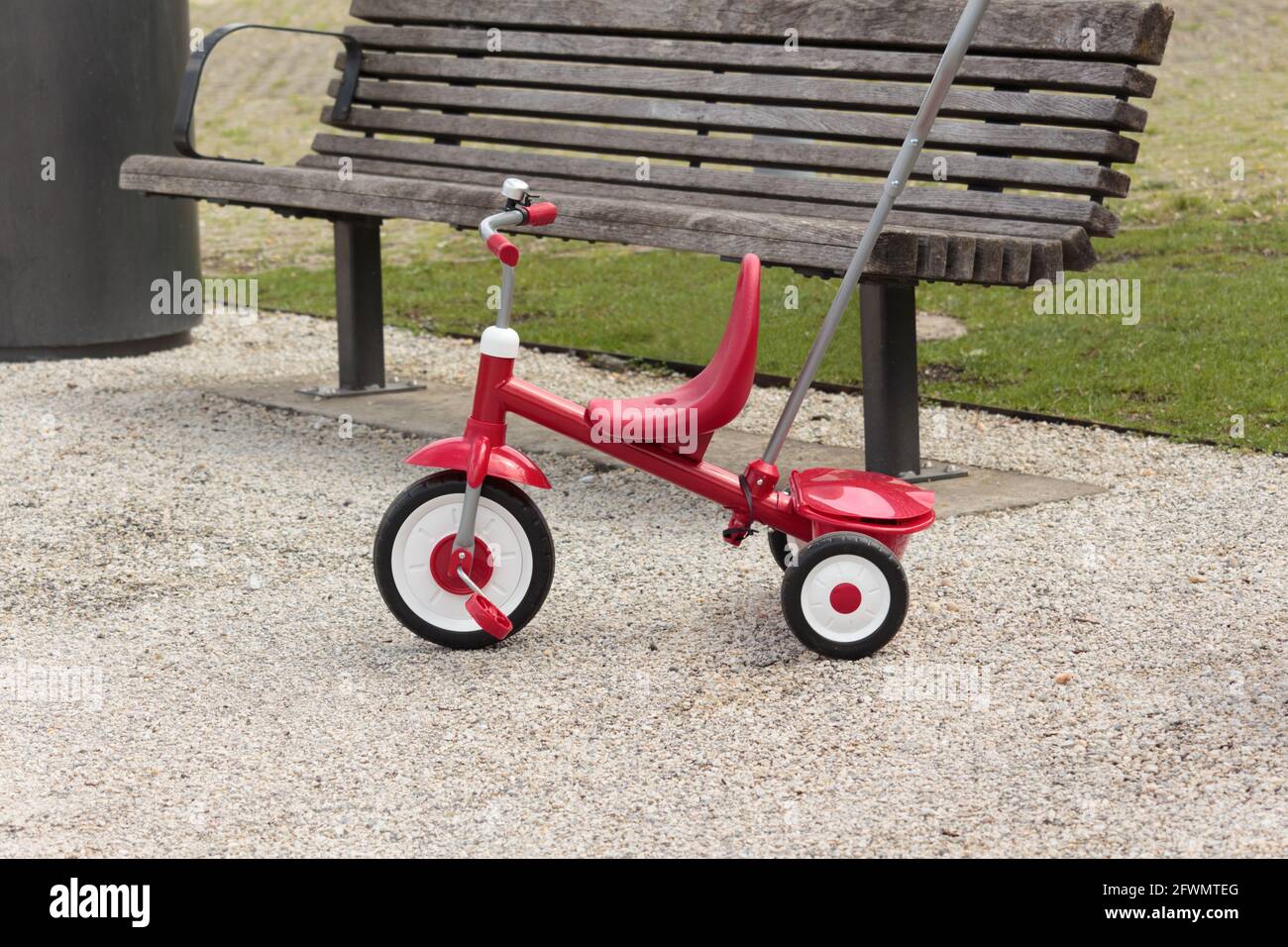 un triciclo rosso vuoto con parcheggiato su una passerella in ghiaia accanto a una panca di parcheggio vuota Foto Stock