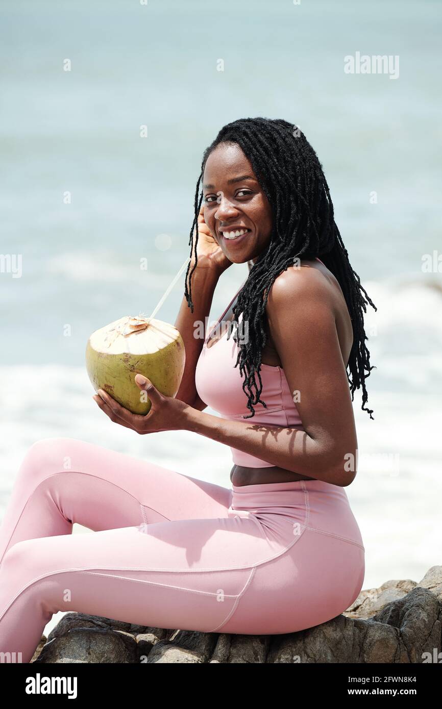 Ritratto di bella giovane donna in abbigliamento sportivo bevendo cocktail di cocco e sorridendo alla macchina fotografica Foto Stock