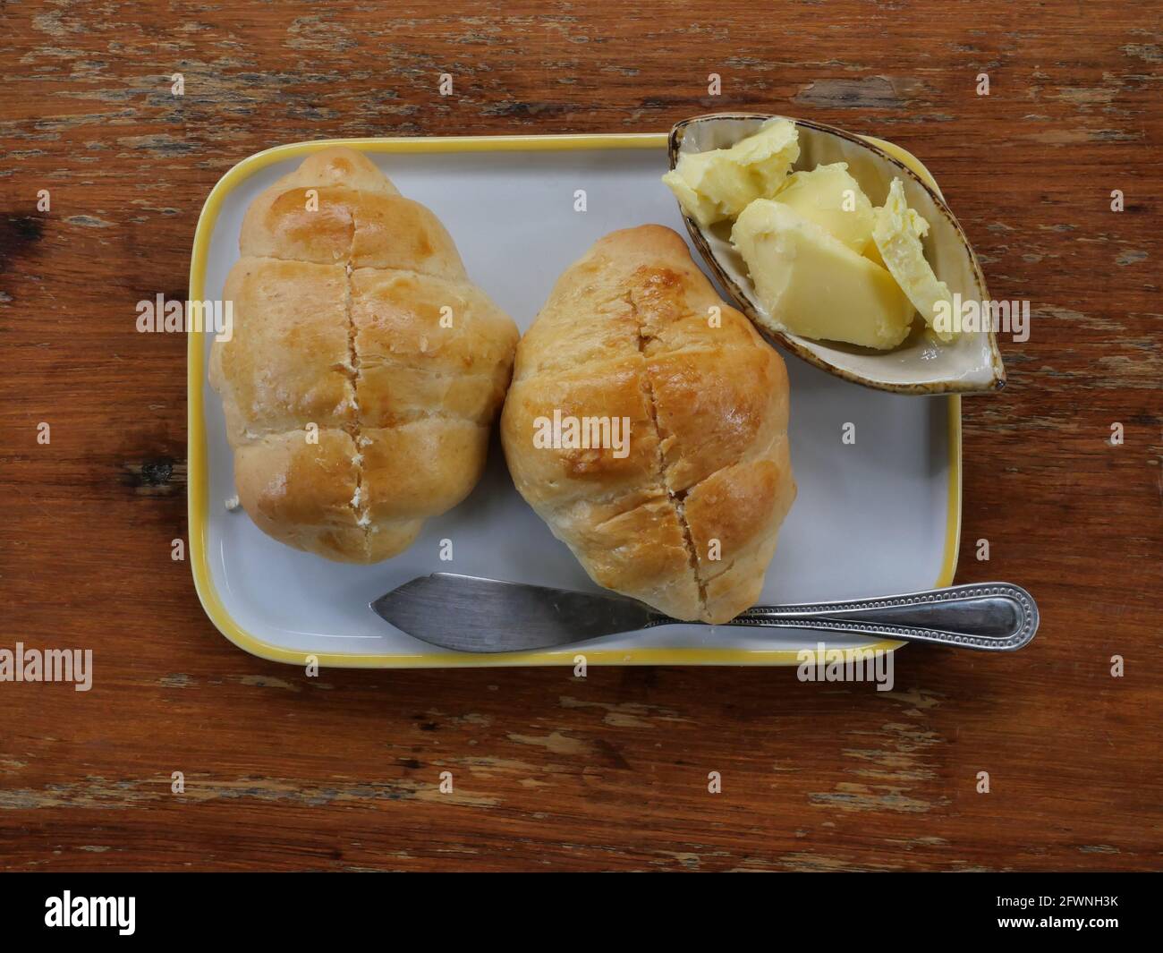 Due croissant freschi su piatto con burro in tazza e coltello, pane francese su tavola di legno marrone Foto Stock