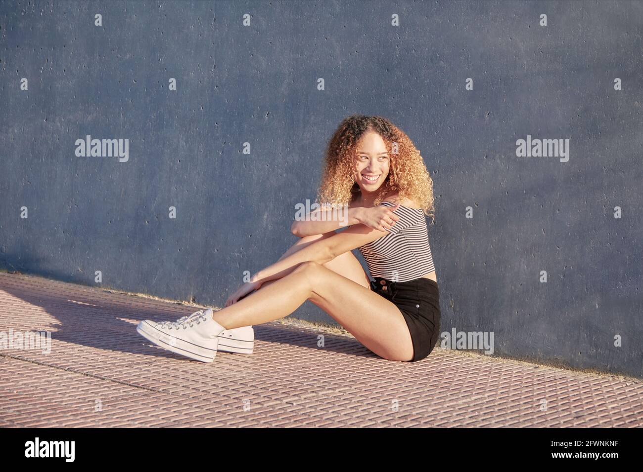 Bella ragazza latina con capelli ricci con pantaloni corti e sneaker bianche, seduta a terra. Ragazza latina sorridente in una giornata di sole. Foto di alta qualità Foto Stock