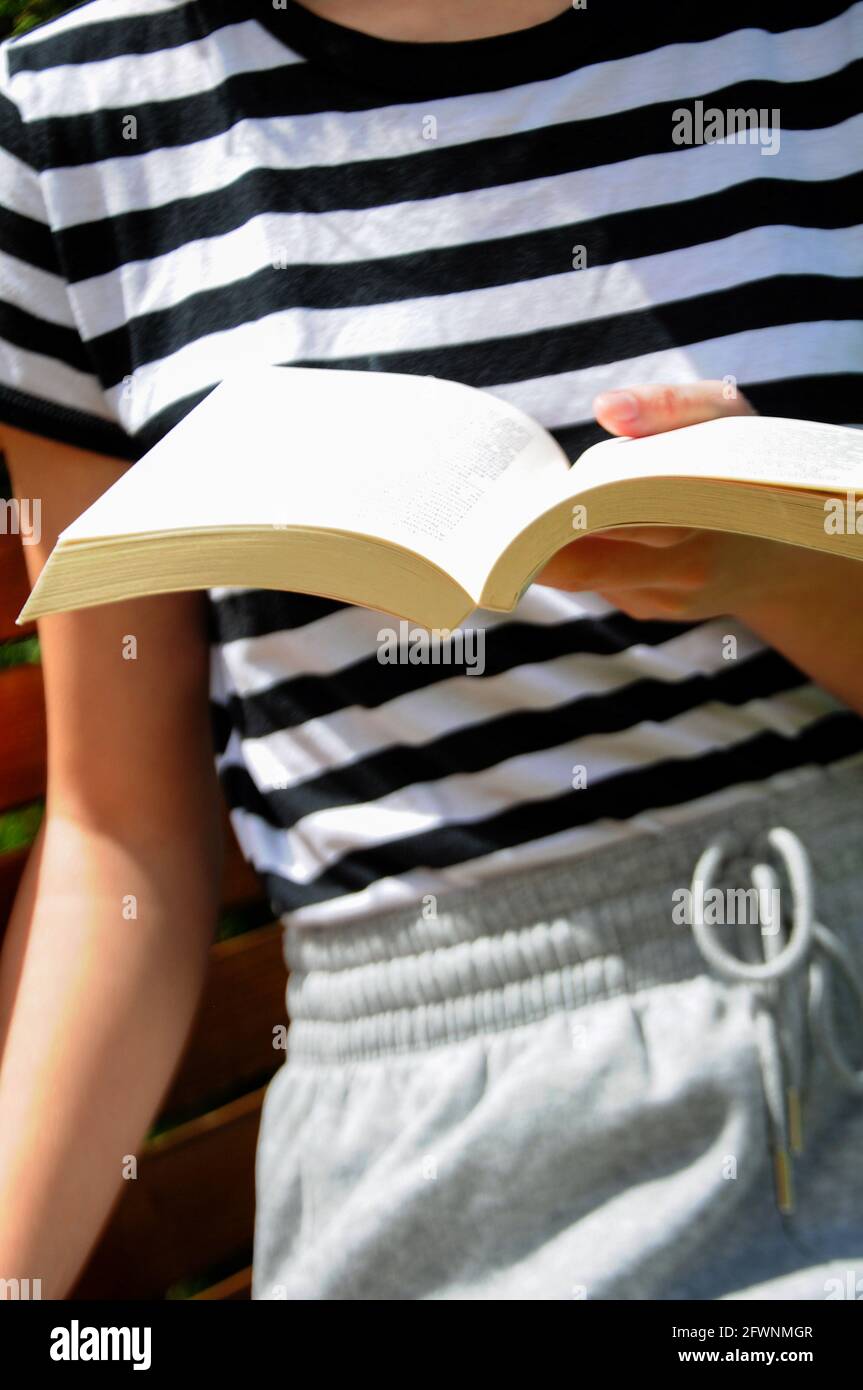 Persona seduta sulla panchina, tenendo un libro bianco e leggendo all'aperto. Tempo libero in giardino. Foto Stock