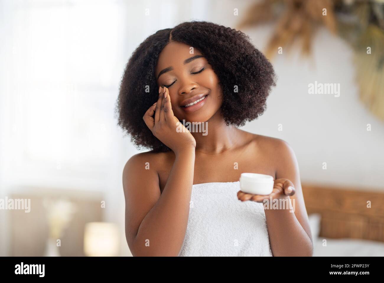 Bella giovane donna nera che applica crema viso dal vaso, indossando asciugamano da bagno, godendo di procedura termale a casa Foto Stock