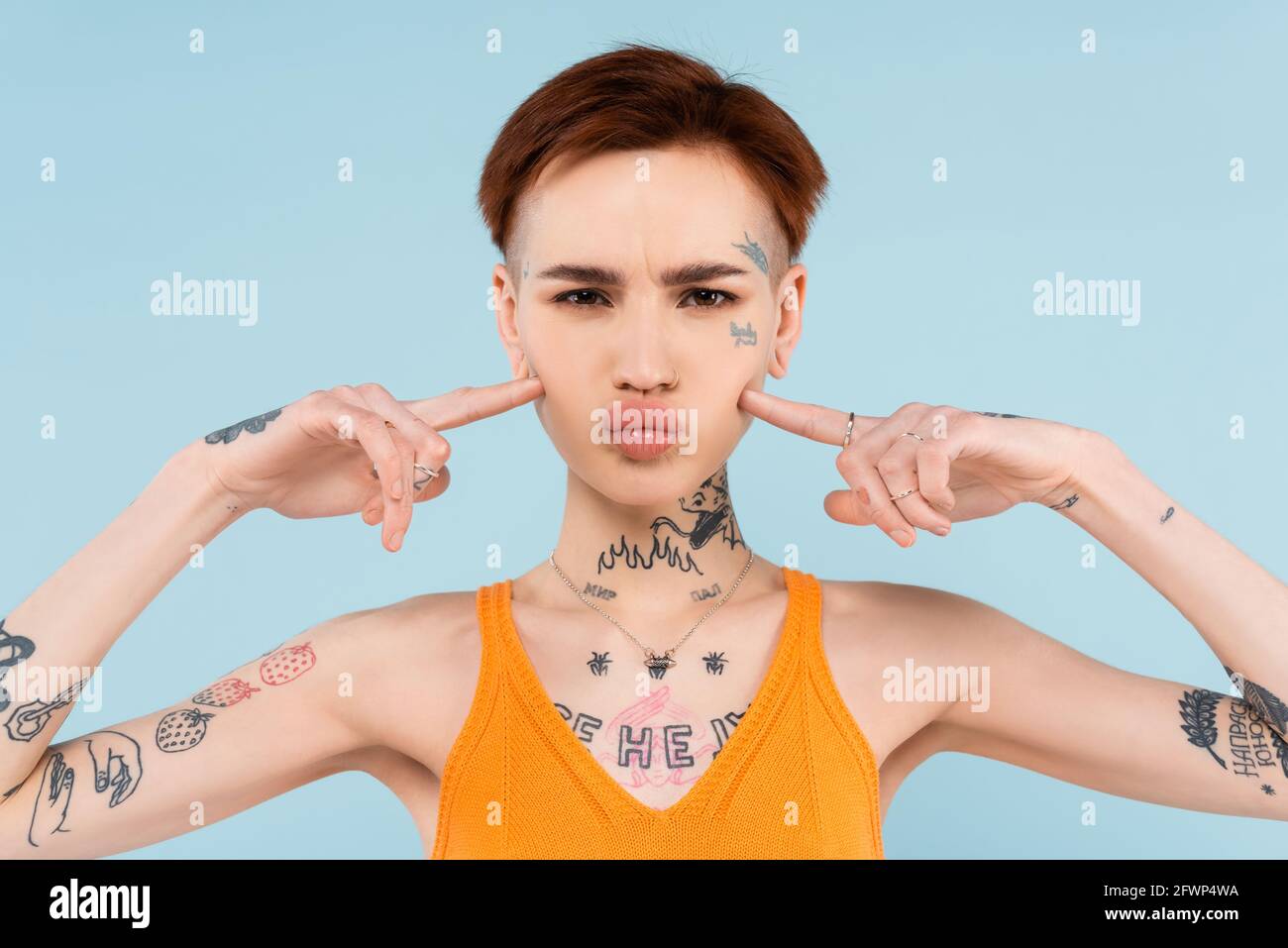 donna con tatuaggi e canotta in maglia arancione che punta guance di puffing isolate su blu Foto Stock