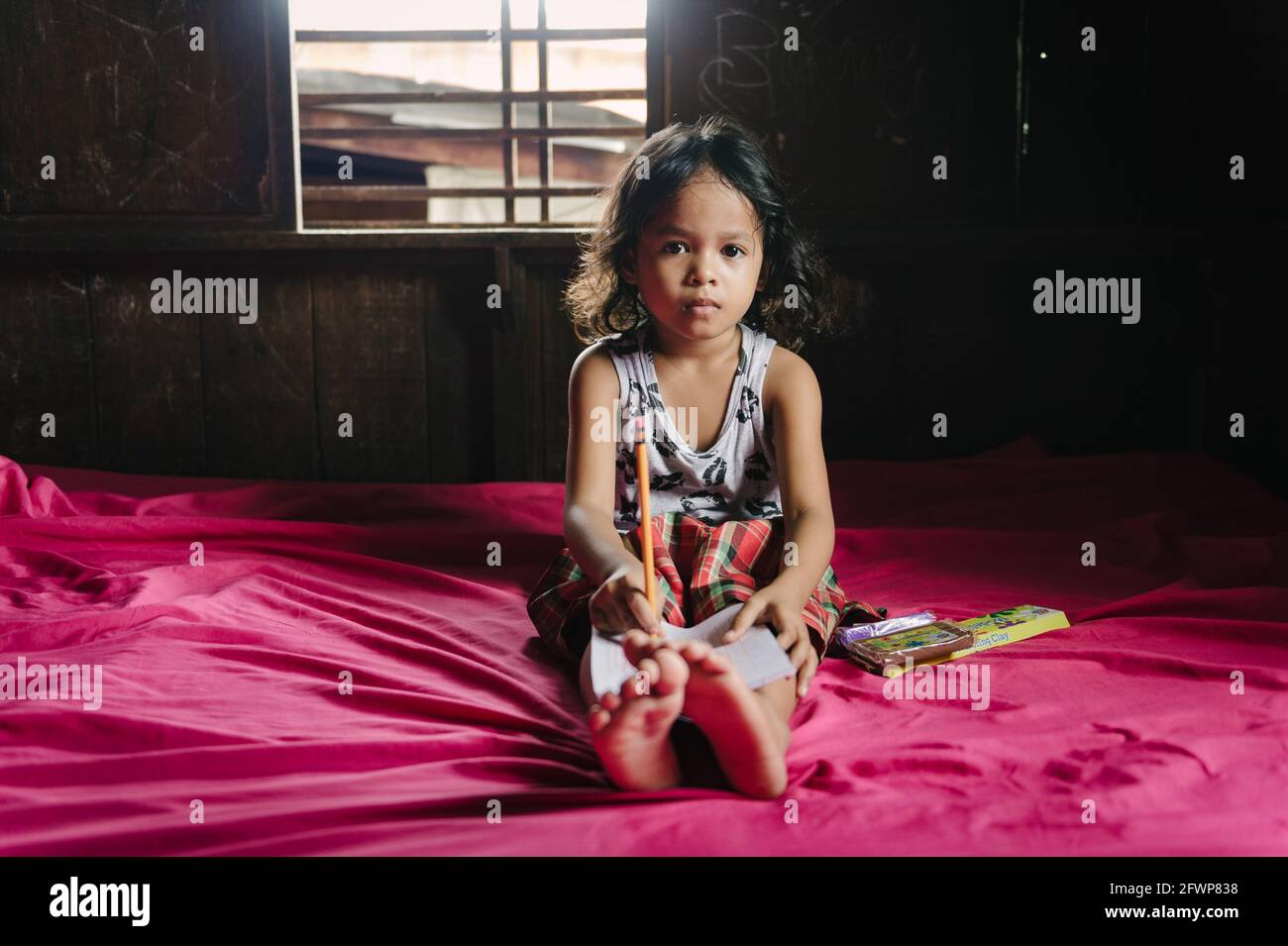 Una giovane studentessa filippina elementare si siede sul letto nella sua piccola casa, facendo i suoi compiti. Foto Stock
