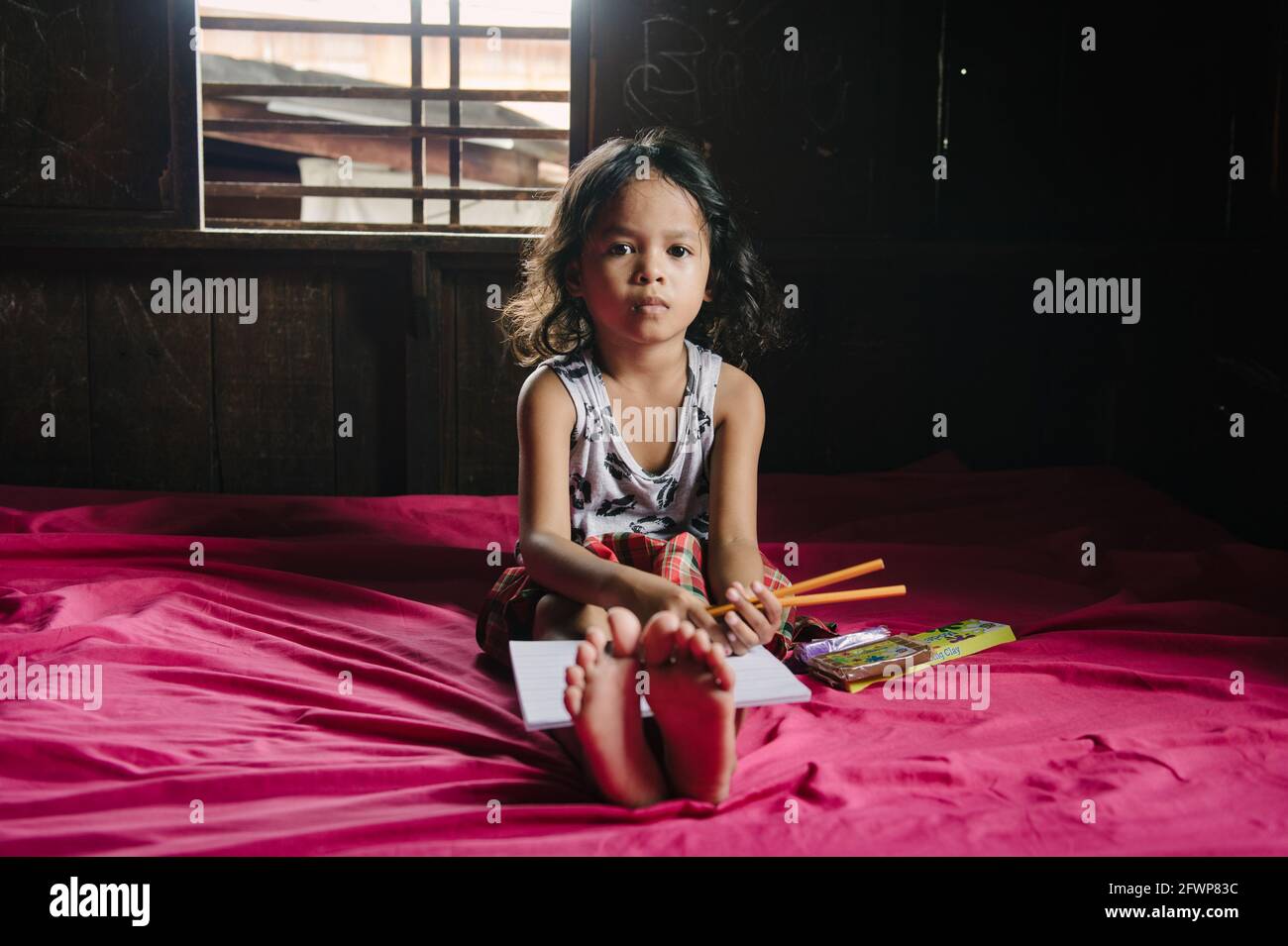 Una giovane studentessa filippina elementare si siede sul letto nella sua piccola casa, facendo i suoi compiti. Foto Stock