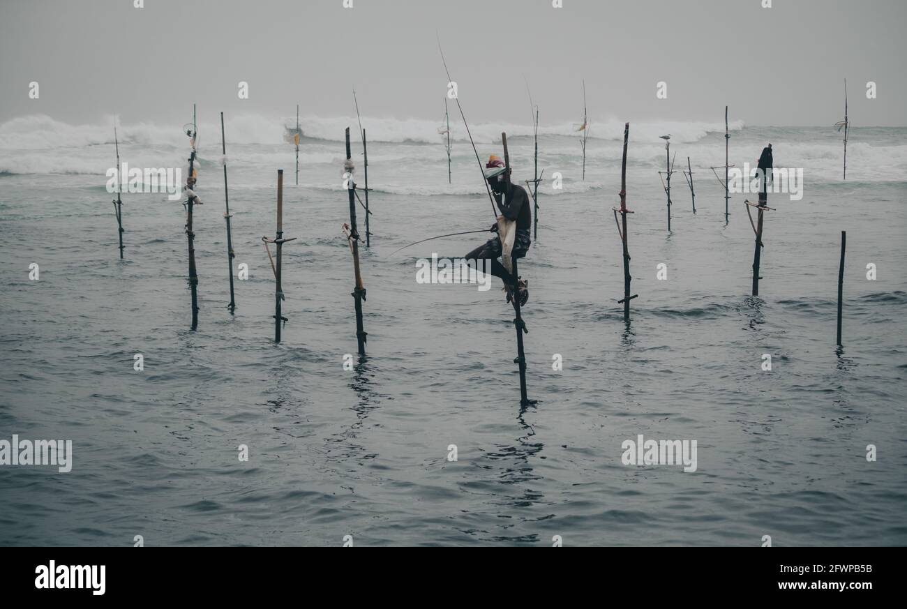 Mirissa, Sri Lanka - 07 26 2020: Pescatori tradizionali di stira che tengono una canna da pesca, e una borsa per la raccolta di pesce, seduti su una croce bastone sopra il Foto Stock