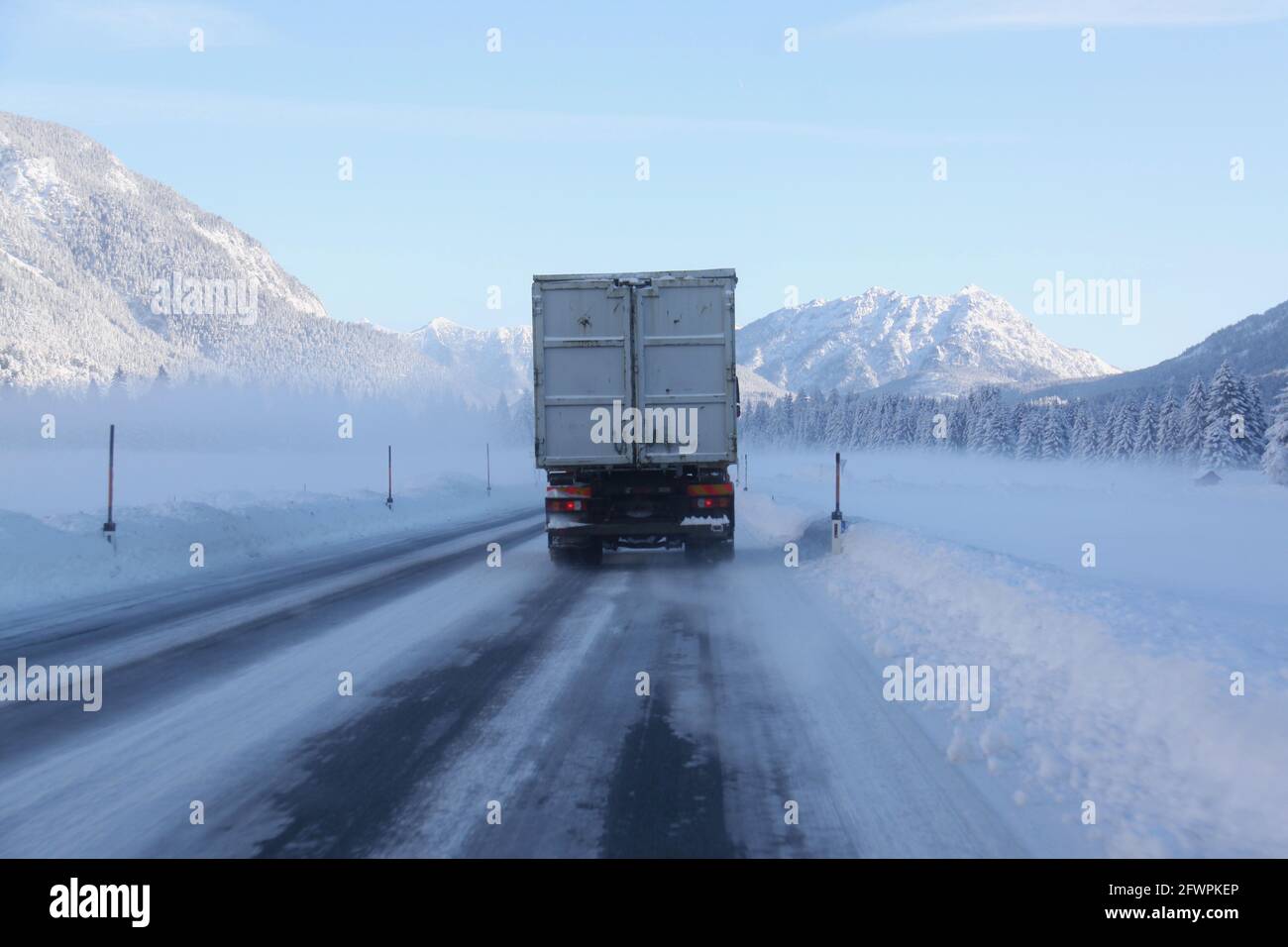 camion bianco che guida su una strada ghiacciata attraverso una neve paesaggio con il sole sulle montagne e nebbia sopra i prati della valle Foto Stock