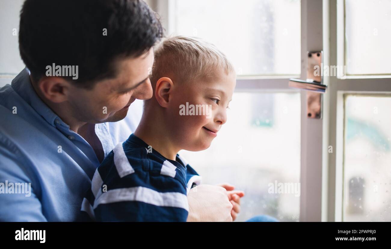 Padre con il figlio della sindrome di Down felice in casa, guardando attraverso la finestra. Foto Stock