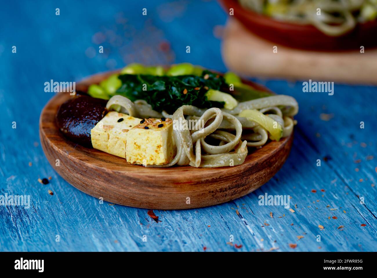 closeup di un piatto di legno, su un tavolo di legno blu, con alcuni dadi di tofu, tagliatelle di farro, fagioli di edamame e kale, e un altro piatto vegano nel backgr Foto Stock