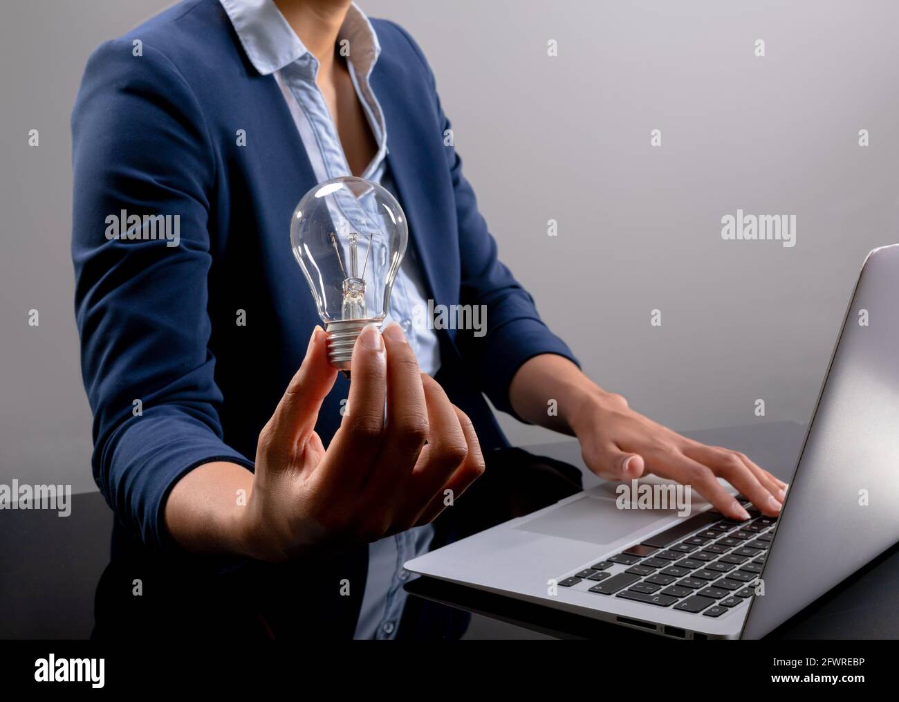 Sezione centrale di donna d'affari che tiene una lampadina usando il laptop su sfondo grigio Foto Stock