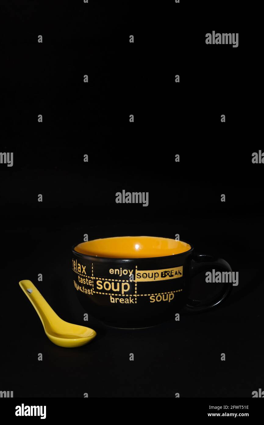 Una zuppa di ceramica o tazza per zuppa con manico e un cucchiaio giallo per zuppa tenuto accanto ad esso con fondo nero piano Foto Stock