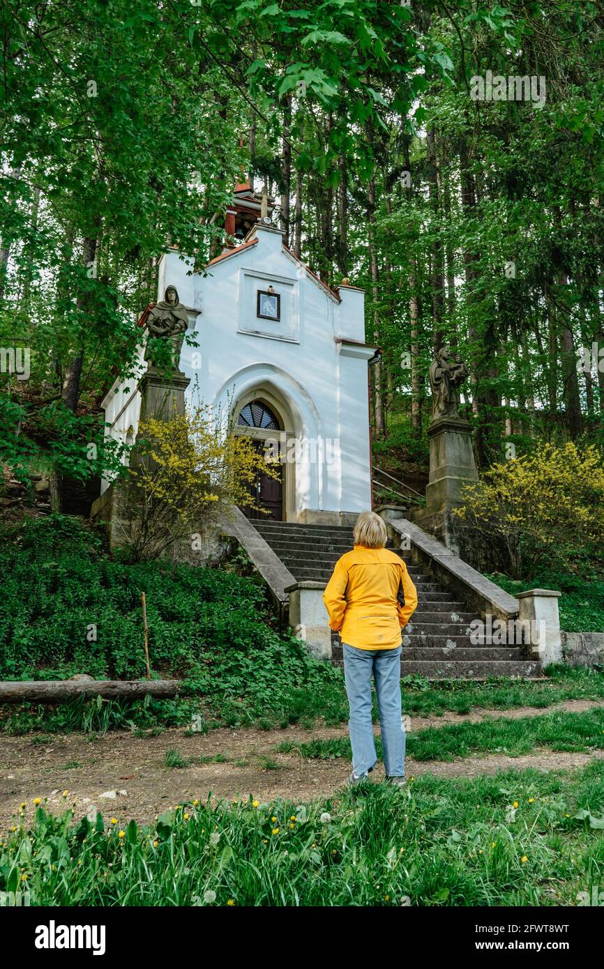 Viaggiatore femminile guardando piccola cappella di nostra Signora di Lourdes, regione di Broumovsko, repubblica Ceca. Chiesa cattolica rurale in campagna primavera. Foto Stock
