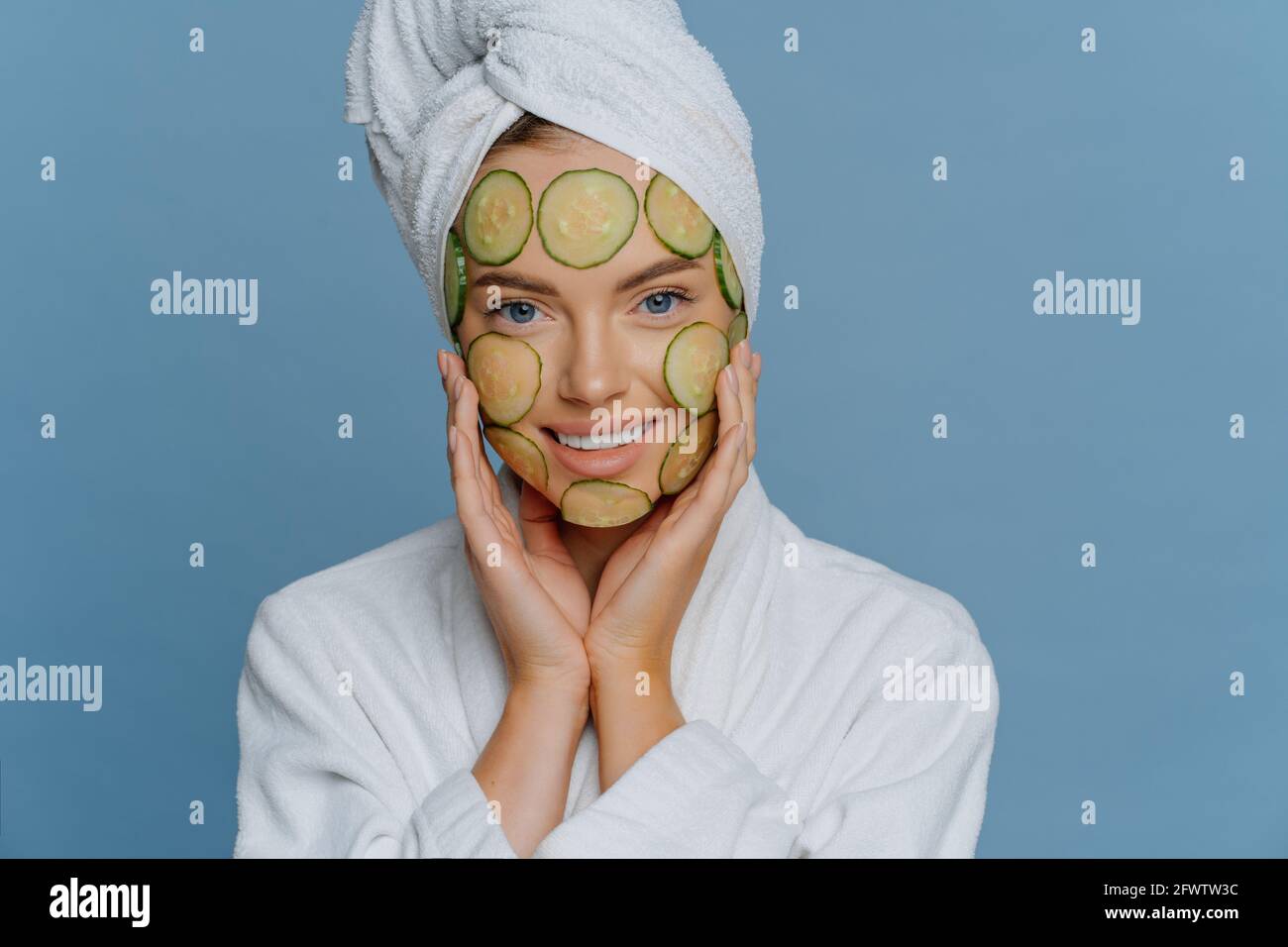 Bella donna applica le fette di cetriolo sul viso ha perfettamente pulito pelle tocca le guance delicatamente vestita in abito da condimento comodo ha asciugamano avvolto su Foto Stock