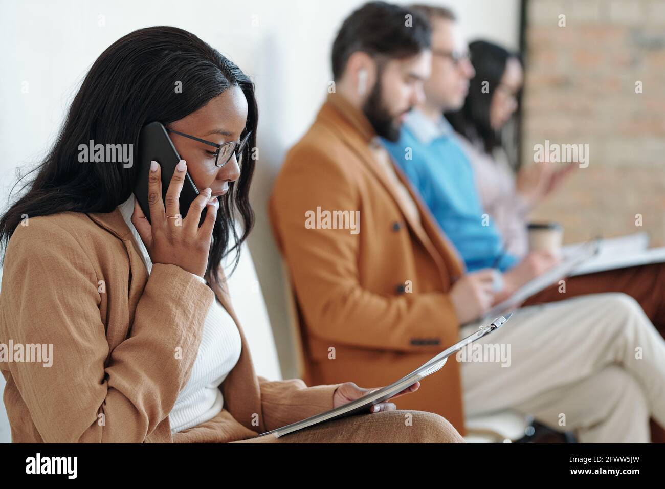 Vista laterale della giovane donna nera seria seduta sulla sedia in corridoio e parlare per telefono mentre si attende il lavoro intervista Foto Stock
