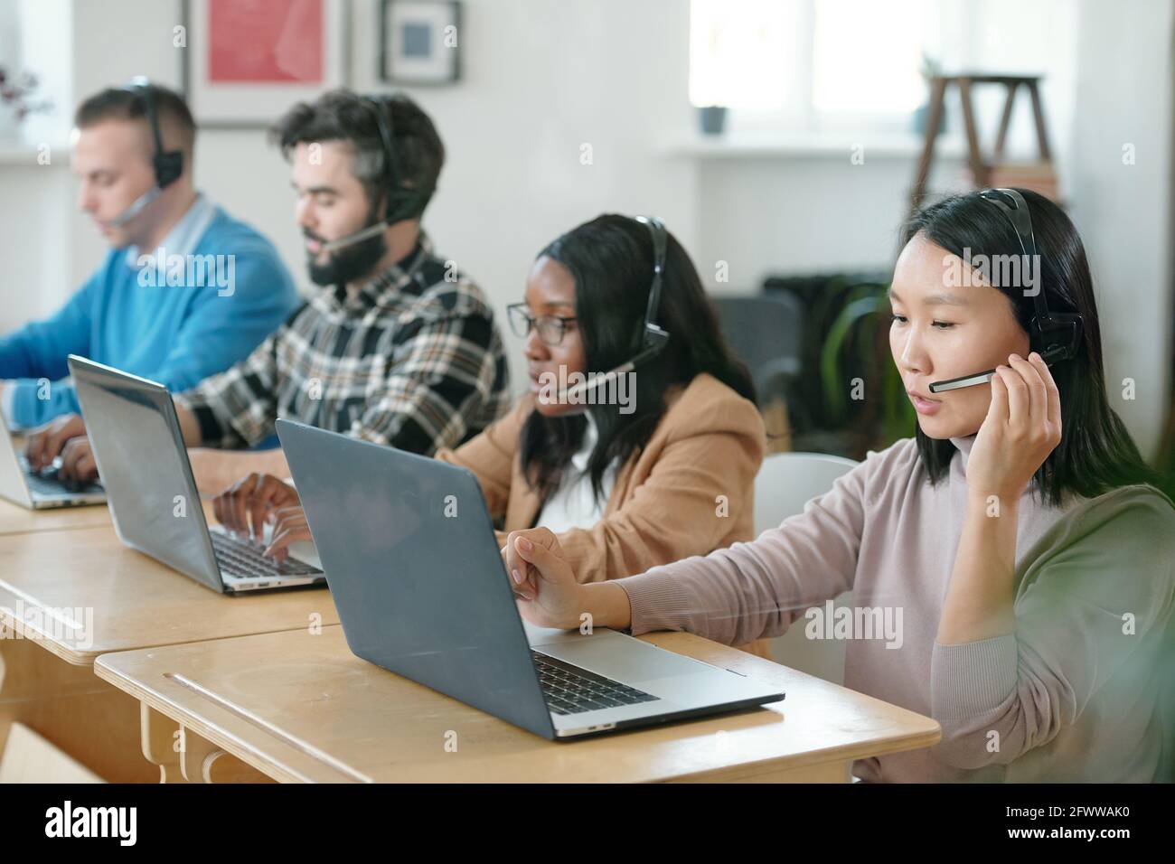 Gruppo di giovani esperti multietnici del servizio clienti che utilizzano cuffie con microfoni e computer portatili durante l'elaborazione delle applicazioni Foto Stock