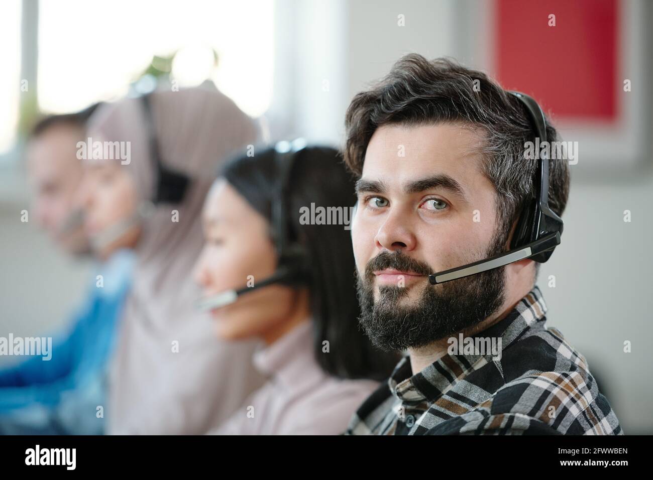 Ritratto dei contenuti di un giovane operatore di assistenza in cuffia wireless con microfono seduto contro colleghi multietnici Foto Stock