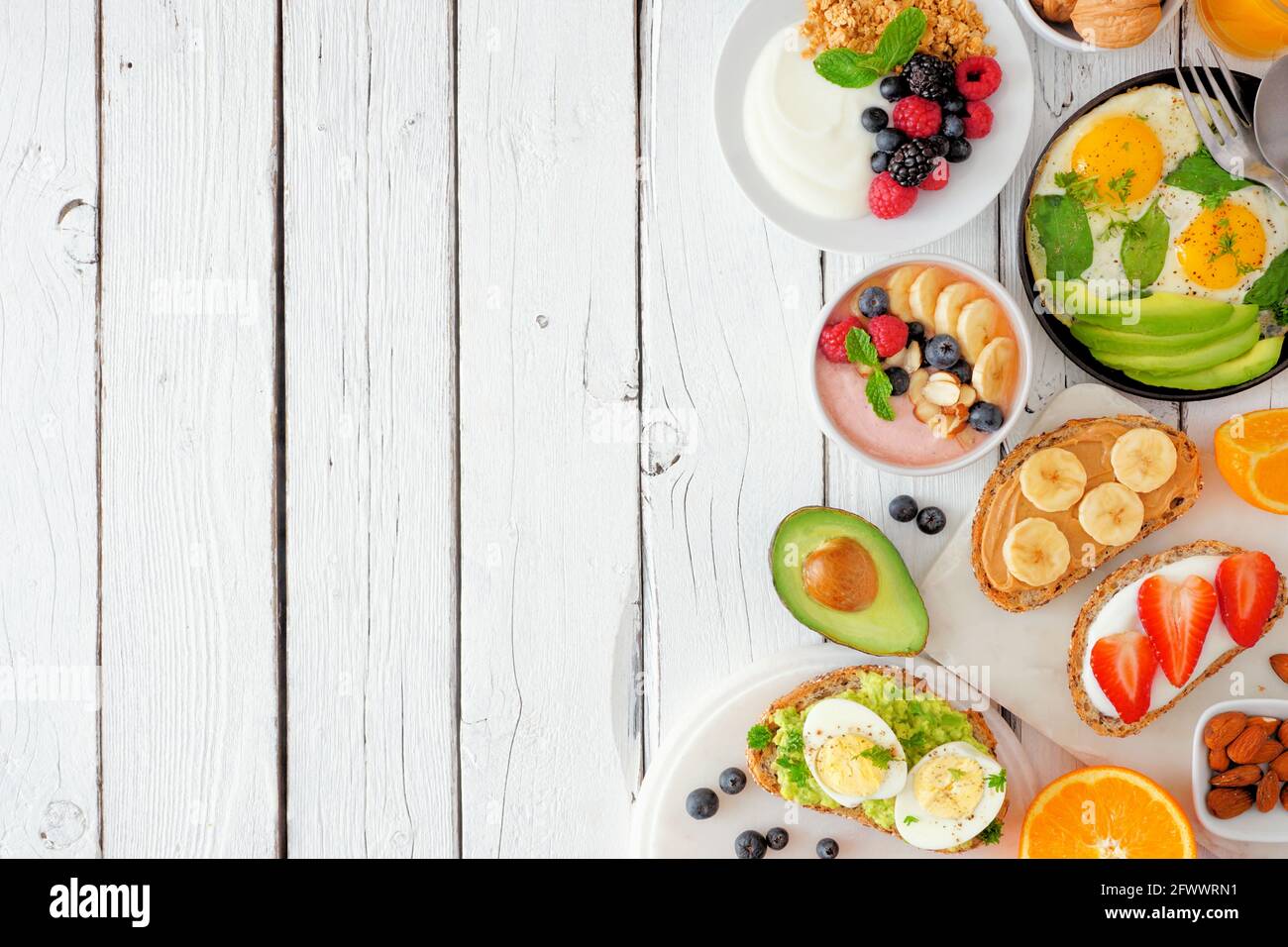 Sana colazione cibo bordo laterale. Tavola con frutta, yogurt, frullati, toast nutrienti e padella d'uovo. Sopra la vista su un backgrou di legno bianco Foto Stock