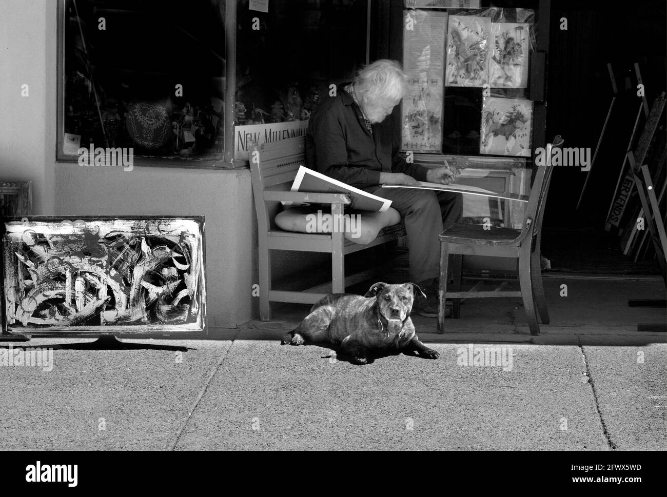 Un artista anziano lavora su un disegno mentre si siede all'ombra davanti ad una galleria d'arte con il suo cane da compagnia al suo fianco a Santa Fe, New Mexico. Foto Stock