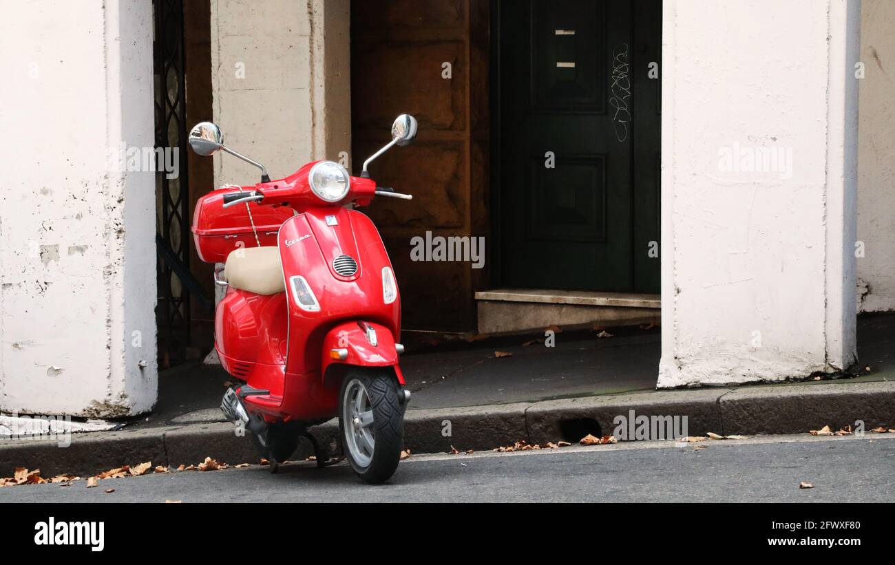 Un simpatico scooter rosso brillante Vespa marchio o moto ciclomotore parcheggiato su una dolce collina con un ambiente rustico e sgargiante edificio della città dietro. Foto Stock