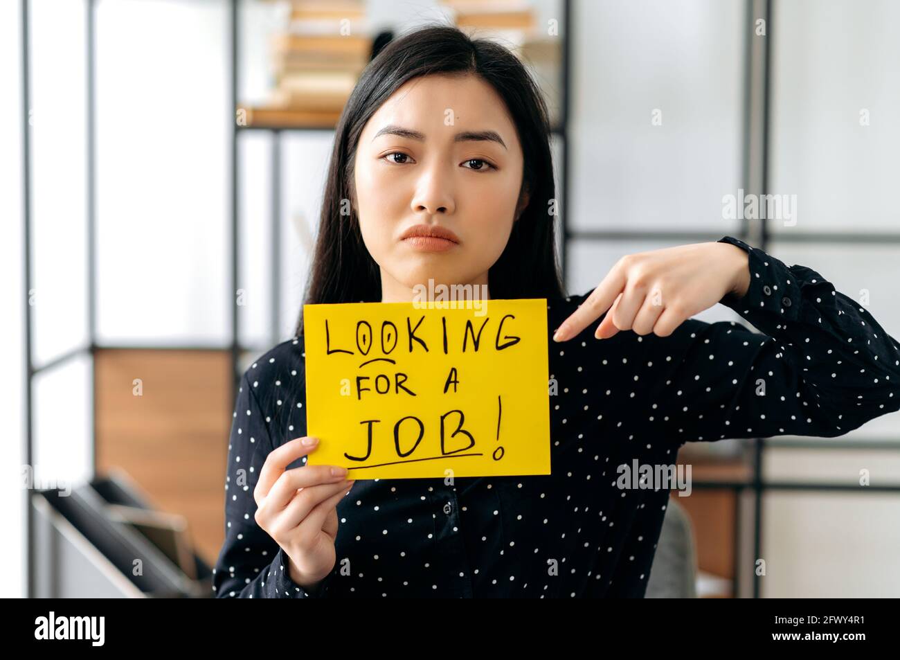 Arrabbiato studente di donna asiatica disoccupata o freelancer, si siede con il segno con l'iscrizione alla ricerca di un lavoro, sperando di ottenere un lavoro di sogni, guardare tristemente la macchina fotografica, puntando il dito all'iscrizione Foto Stock