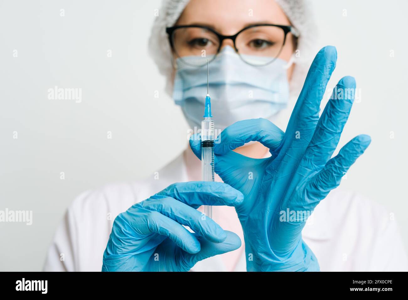 Medico donna o scienziato in bianco camice blu, guanti, cappuccio verde e la maschera contiene una siringa in mani su sfondo bianco. Ella è pronta a dare Foto Stock