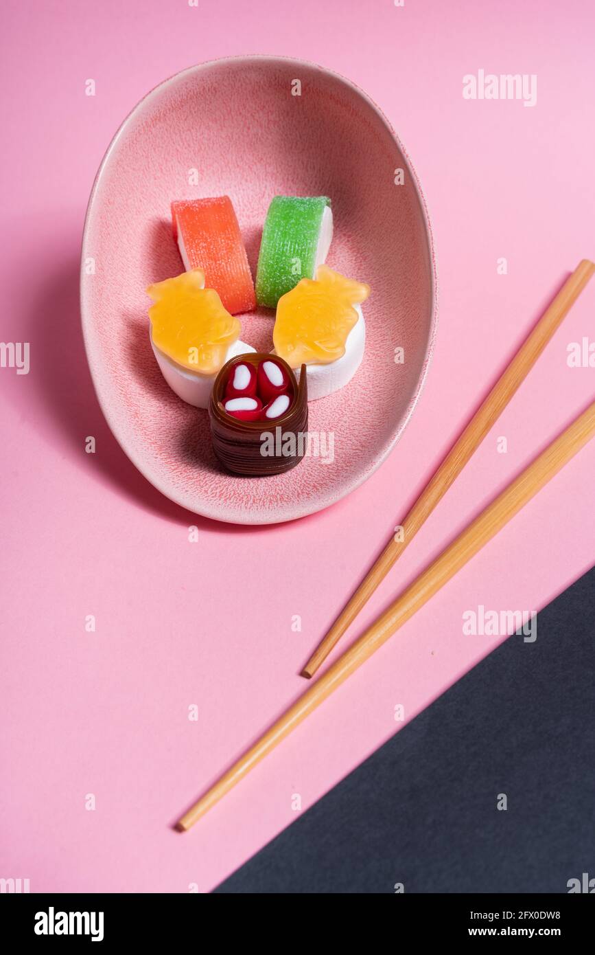 Vista dall'alto di appetitosi sushi dolci colorati serviti su ceramica piatto con bastoncini di legno posti su sfondo rosa e nero Foto Stock