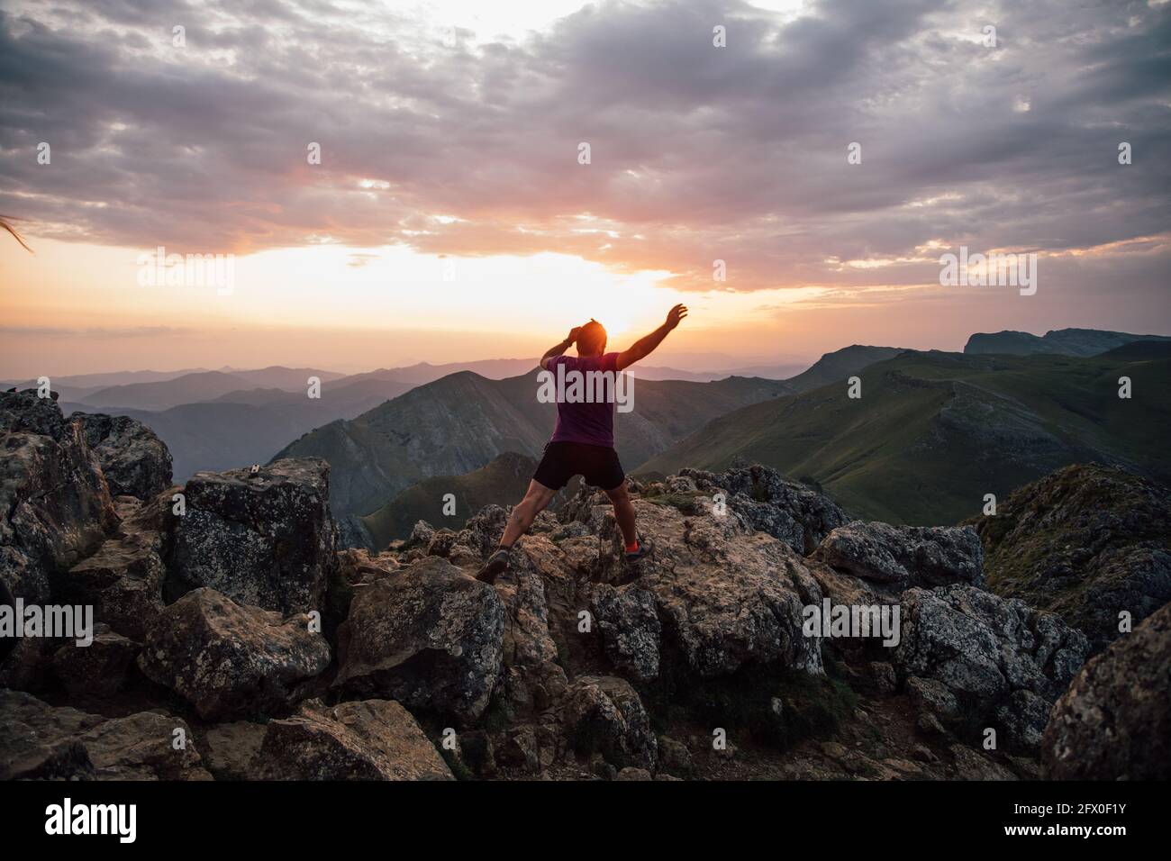 Vista posteriore dell'uomo che salpa su massi di alta cima di cresta di montagna sotto cielo nuvoloso al tramonto Foto Stock