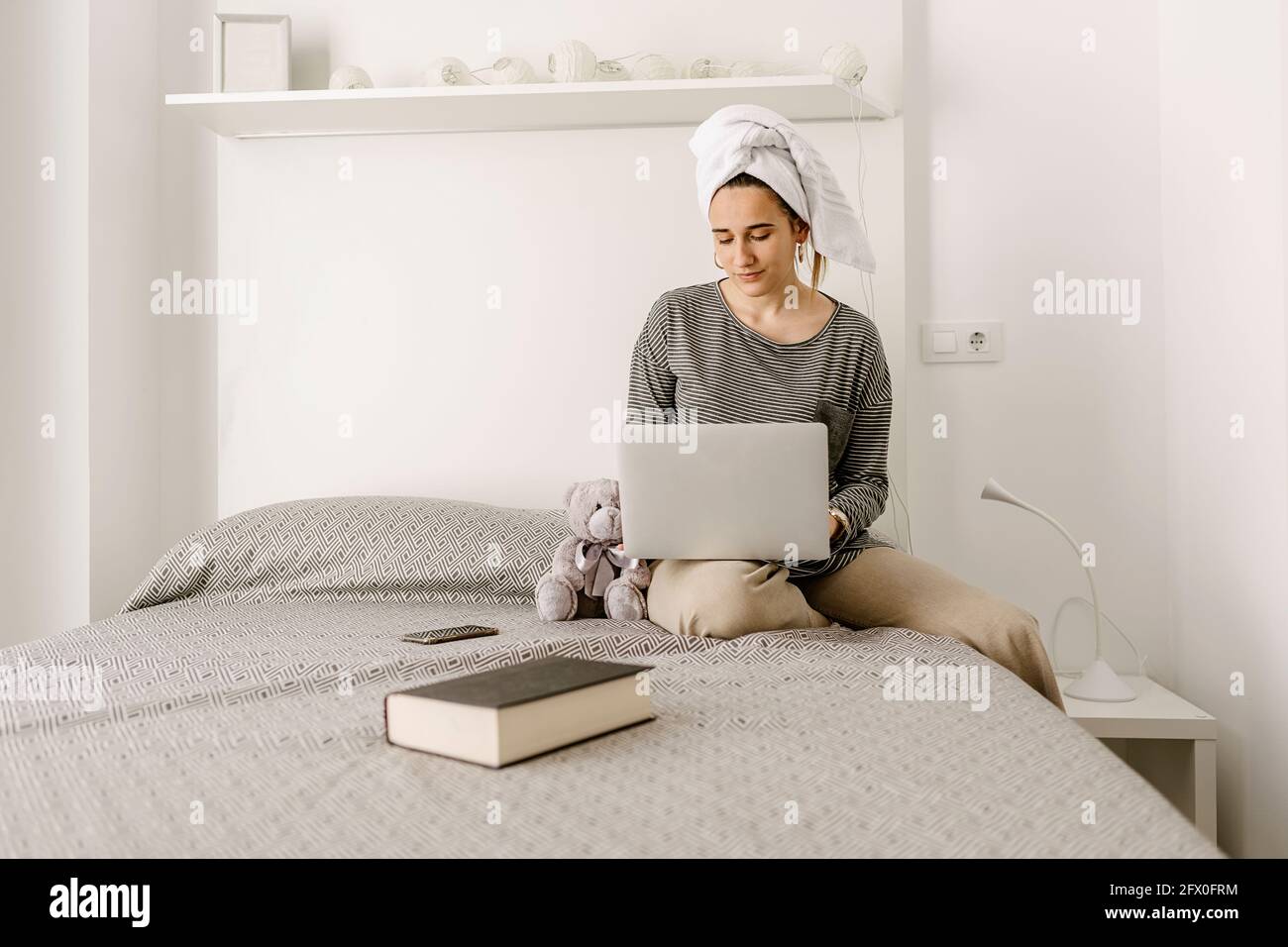 Giovane donna indipendente in abiti e asciugamano casual mentre si lavora a distanza sul netbook mentre si è seduti comodamente letto a casa Foto Stock
