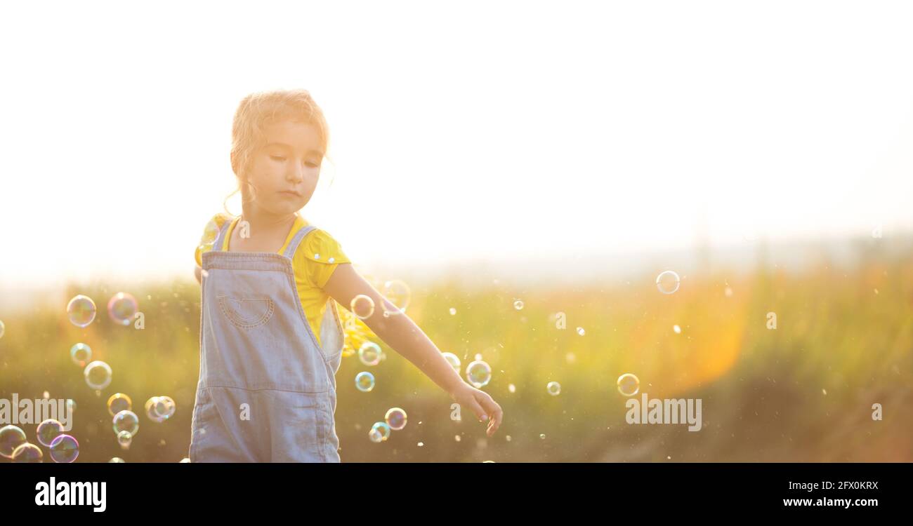 Una ragazza in un jumpsuit denim soffia bolle di sapone in estate in un campo al tramonto. Giornata internazionale dei bambini, Happy child, attività all'aperto. Summe Foto Stock