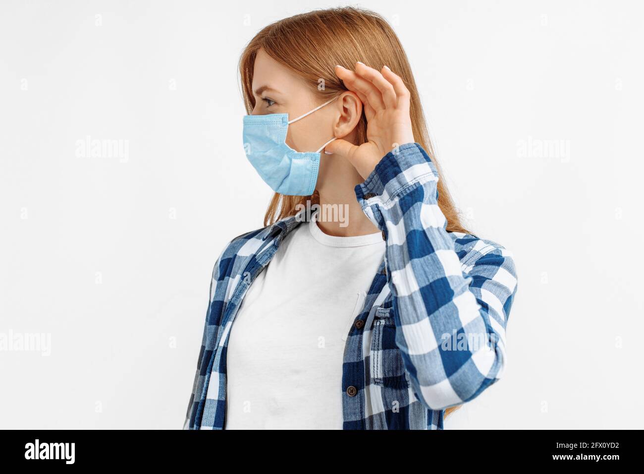 Giovane bella donna che indossa una maschera protettiva da virus che tengono la mano sopra l'orecchio ascoltando voci o pettegolezzi, su sfondo bianco isolato Foto Stock