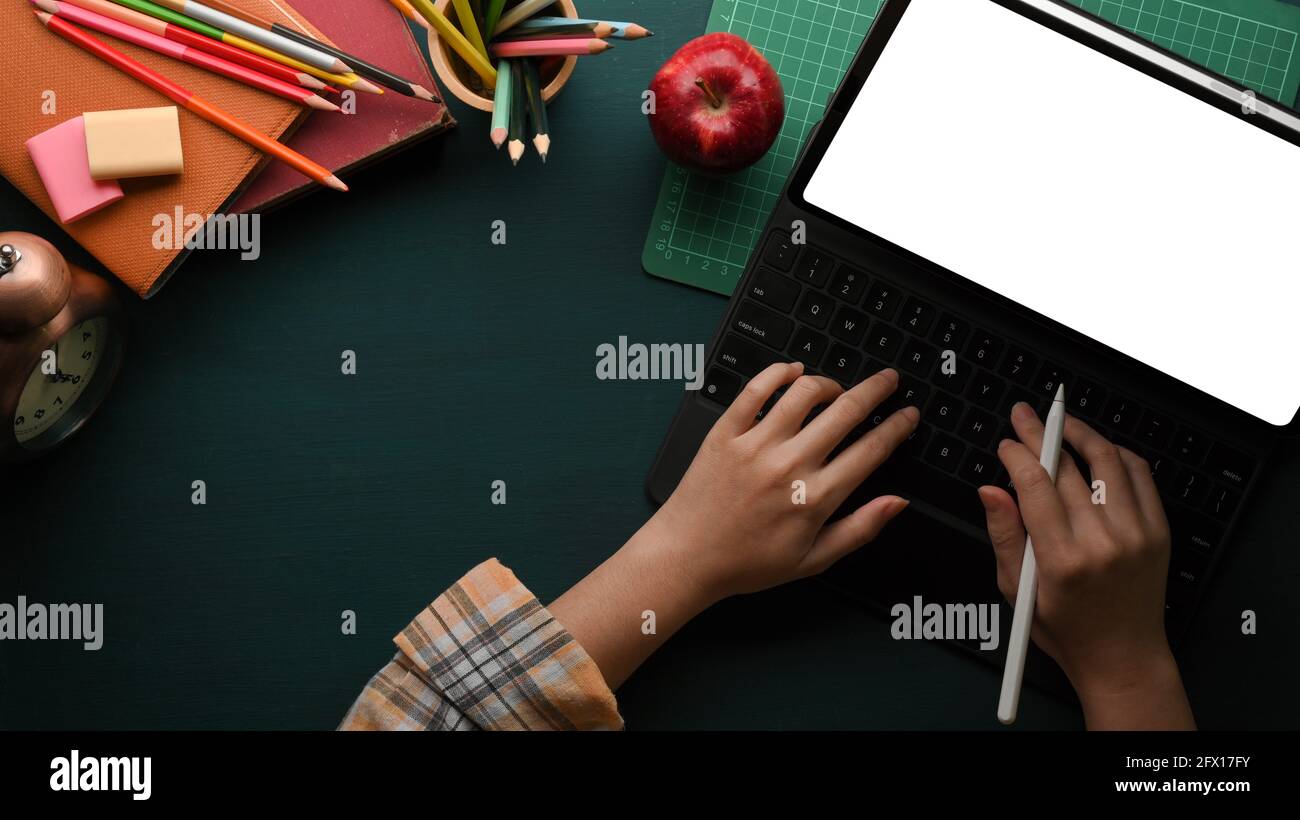 Vista dall'alto della digitazione delle mani femminili sulla tastiera del tablet sul tavolo di studio di colore verde scuro con elementi scolastici, tracciato di ritaglio Foto Stock