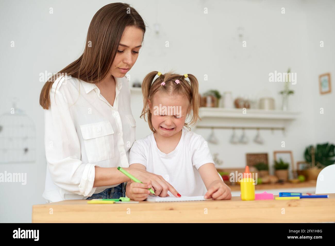 Madre gentile che abbraccia e conforta piccola figlia piangente che ha difficoltà con i compiti mentre sedendo al tavolo e facendo l'esercitazione dentro copybook in Foto Stock