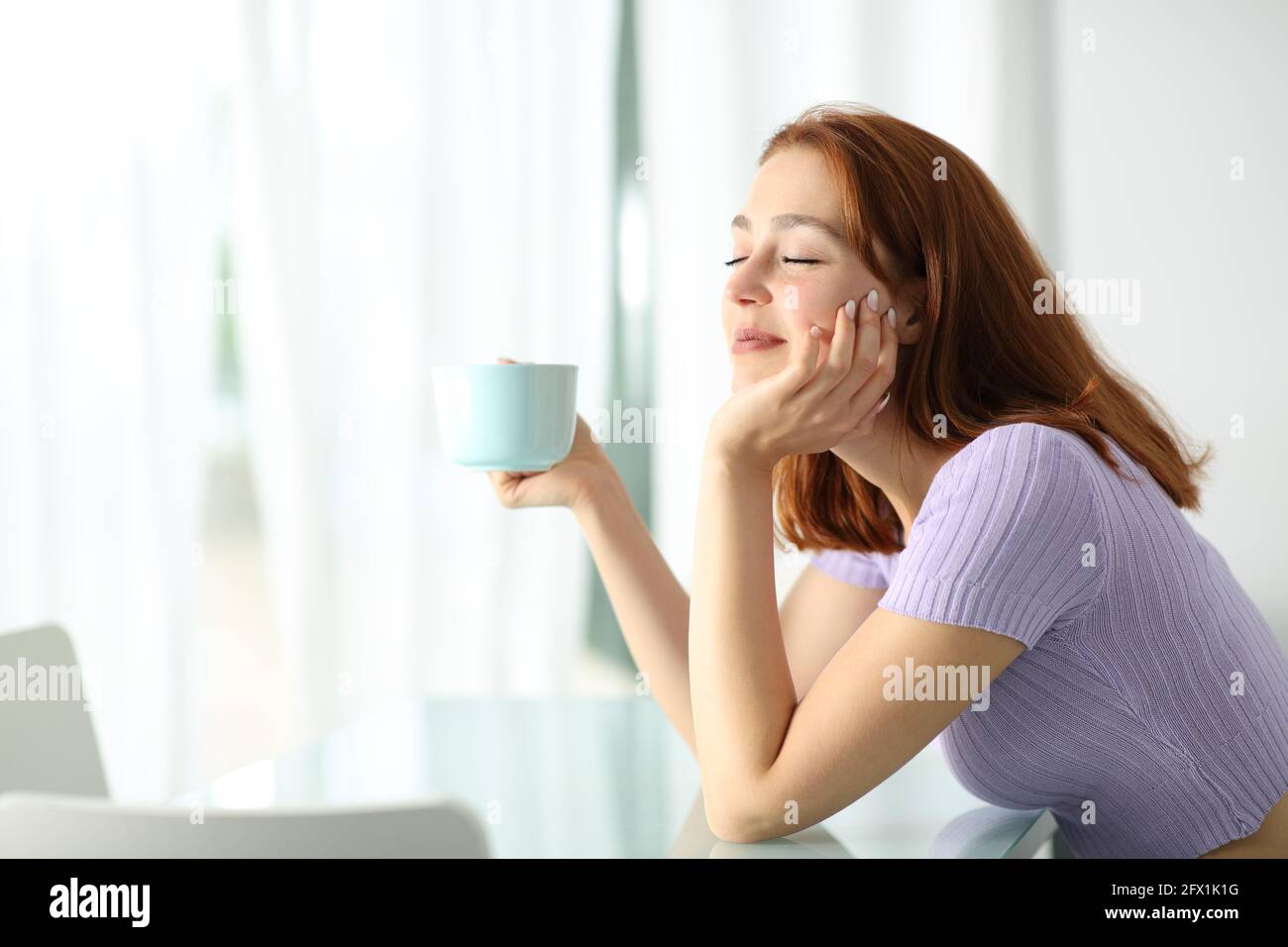 Donna felice che tiene la tazza di caffè con gli occhi chiusi che riposano dentro interno dell'appartamento Foto Stock