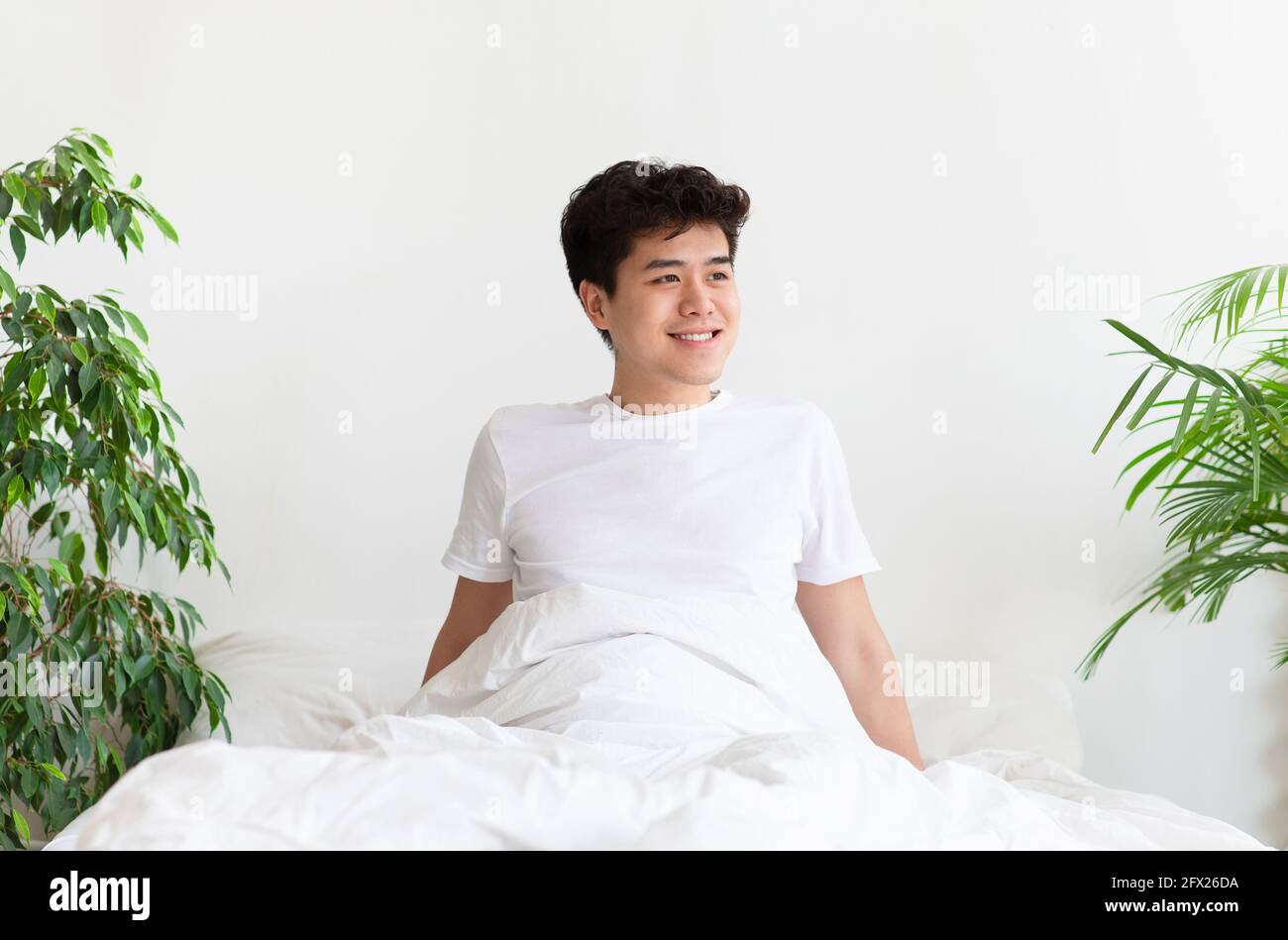 Felice giovane asiatico maschio si sveglia, si siede sul letto e guarda verso spazio vuoto Foto Stock