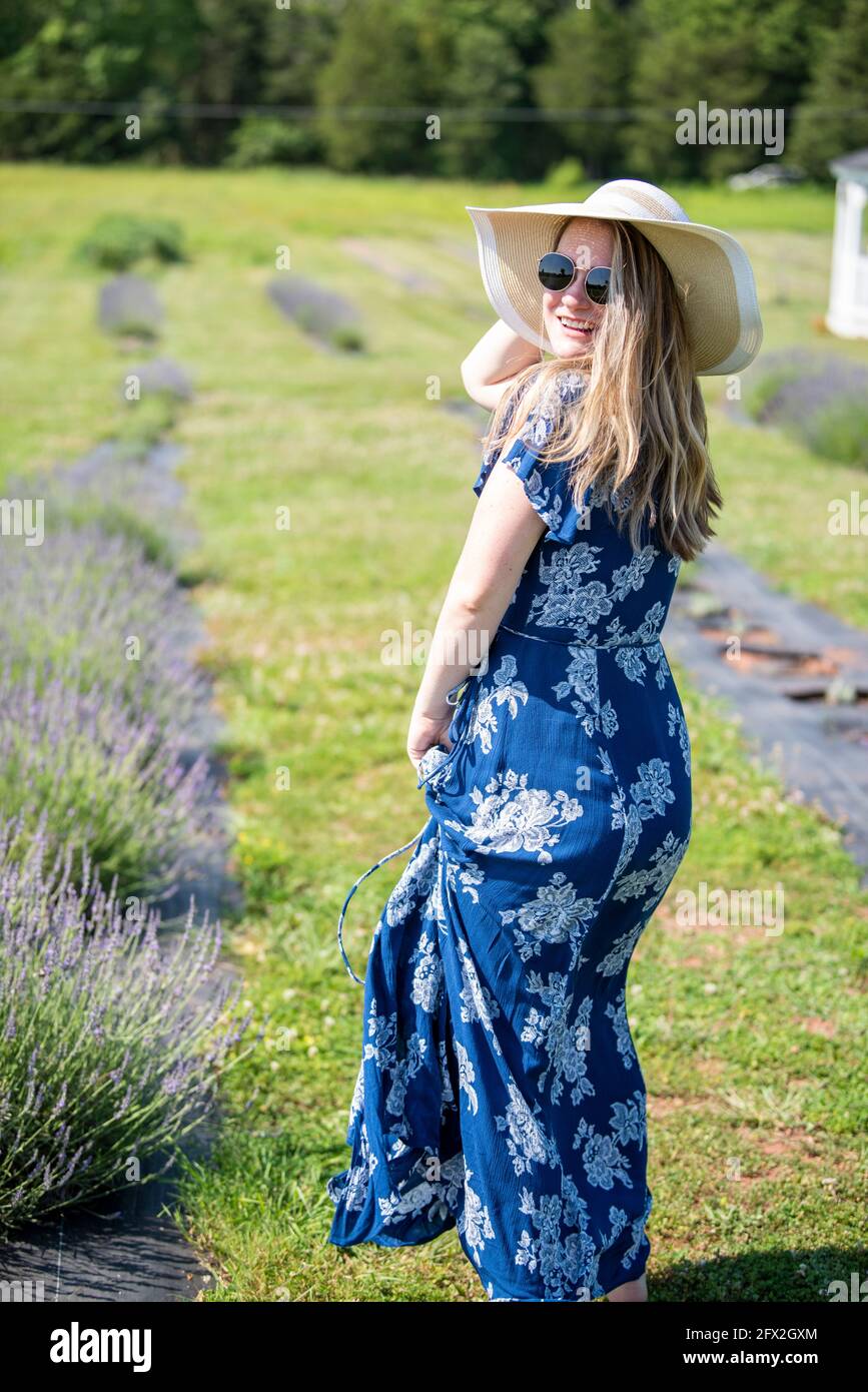 Una donna caucasica che indossa occhiali da sole e un vestito blu cammina in un campo di lavanda fresca. I suoi capelli e vestito soffiano nel vento. Prato in fiore, conto Foto Stock