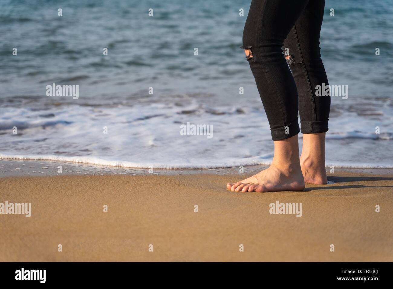 Uomo irriconoscibile in riva al mare. Indossa jeans neri con le maniche arrotolate ed è a piedi nudi. È una giornata di sole. Foto Stock