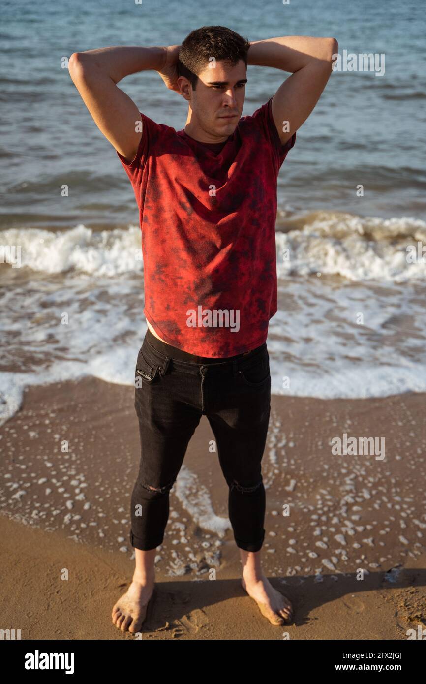 Uomo in posa in mare in una giornata di sole. Caucasica e capelli scuri.  Indossa una t-shirt a maniche corte e pantaloni lunghi arrotolati. Il mare  è calmo. H Foto stock -