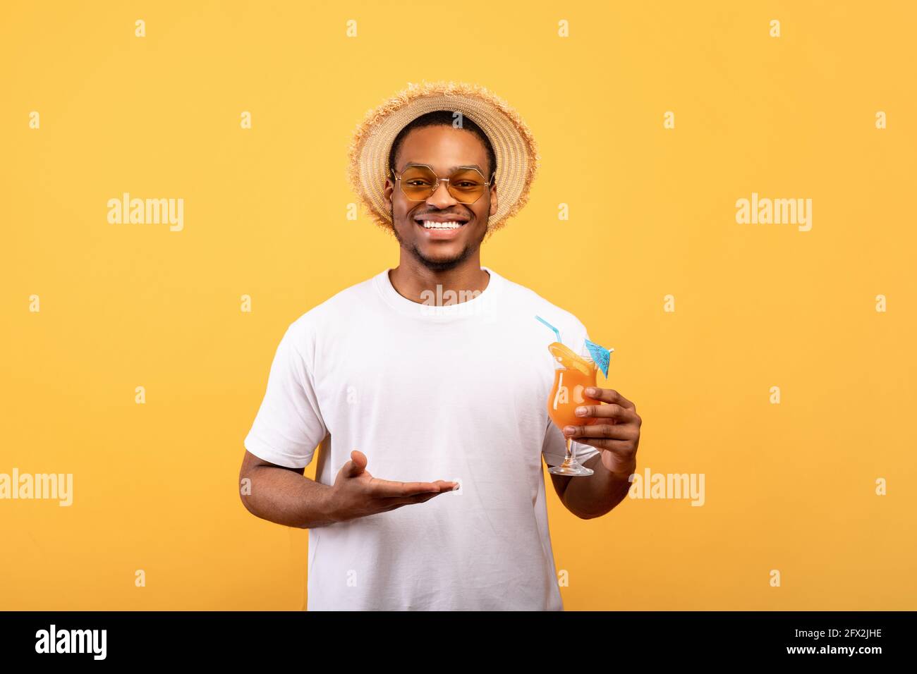 Ragazzo nero millenario che mostra un bicchiere di cocktail tropicale yummy sfondo giallo studio Foto Stock