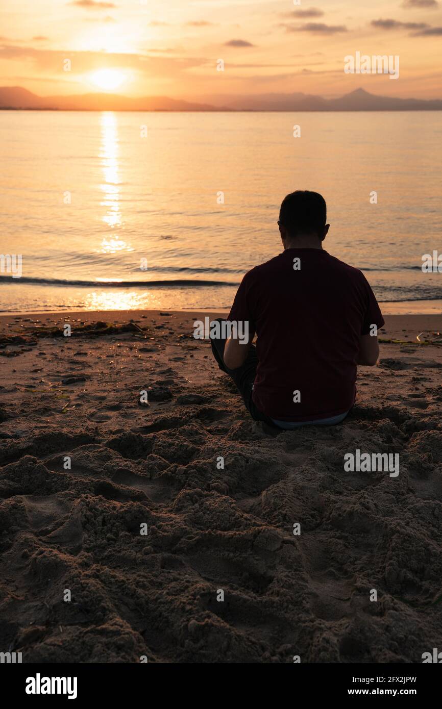 Uomo seduto in riva al mare guardando il tramonto. È seduto sulla sabbia. Il mare è calmo. Mediterraneo, Denia Foto Stock