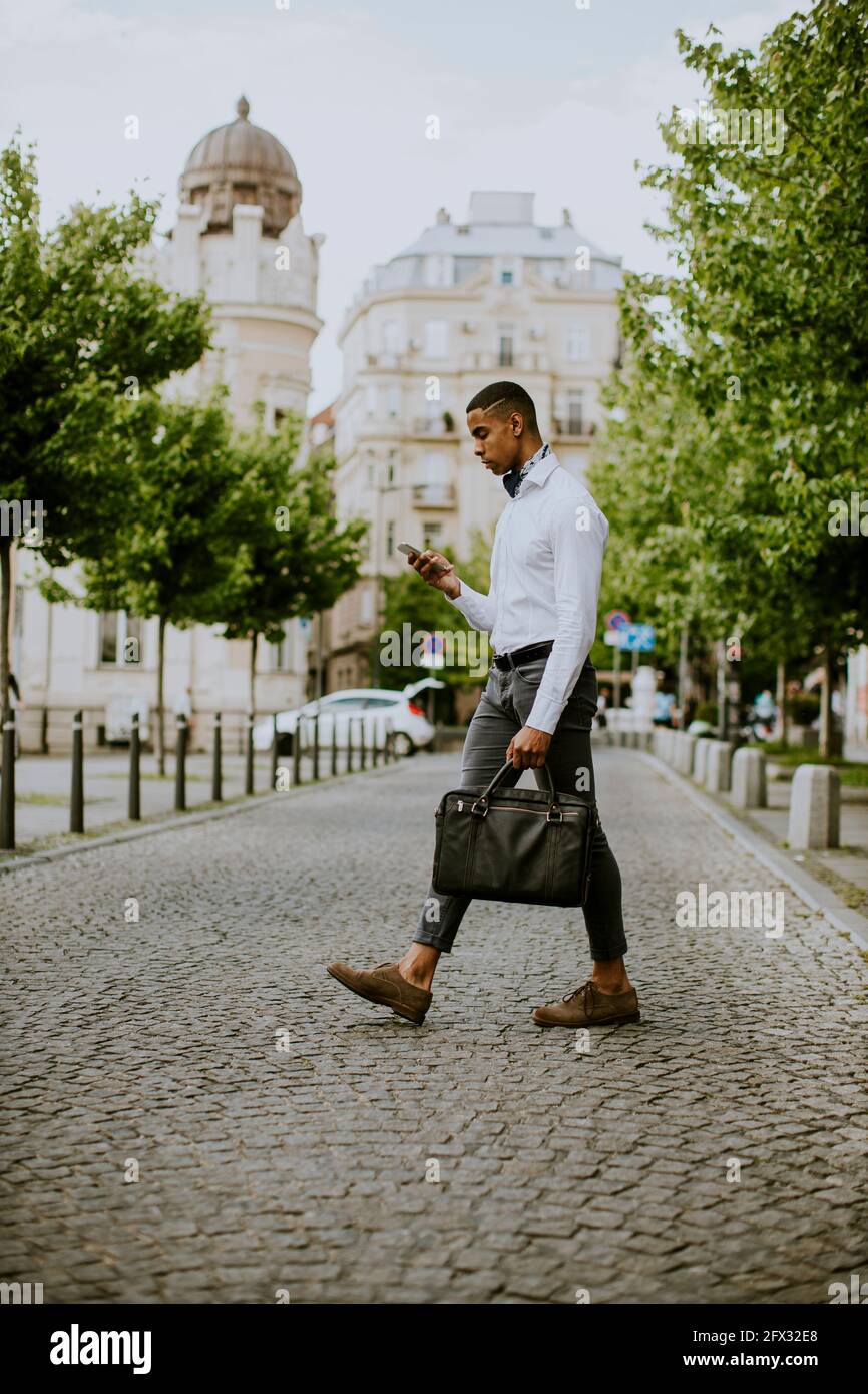 Bel giovane uomo d'affari afro-americano che usa un telefono cellulare mentre ceossing una strada Foto Stock