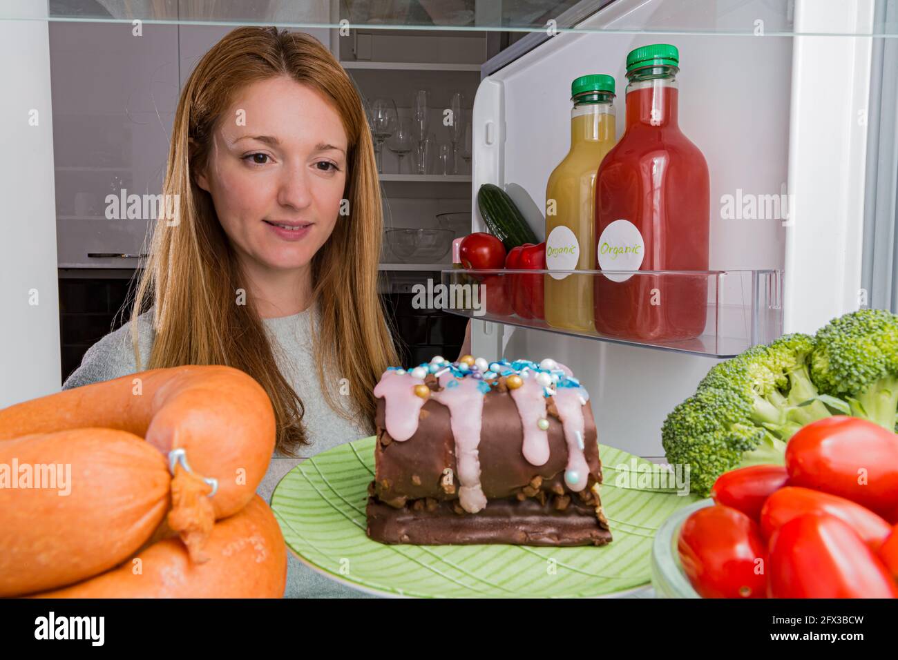 Frigorifero aperto dall'interno, ripiani in vetro con diversi alimenti. Alimentazione malsana, concetto di zucchero alimentare. Giovane donna che guarda la torta al cioccolato Foto Stock