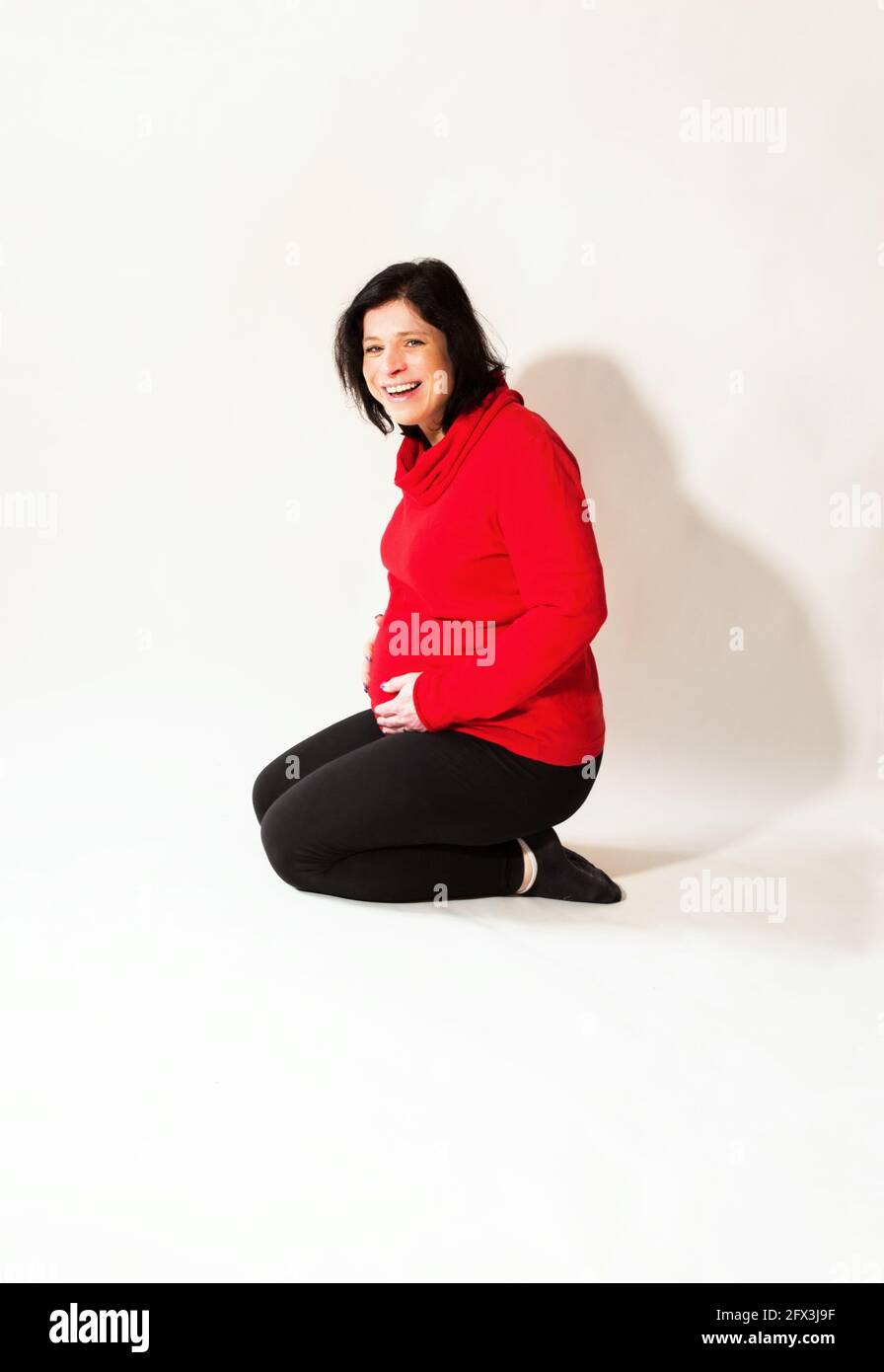 Giovane attraente donna incinta con il sorriso malizioso inginocchia e tiene per la pancia. Su sfondo bianco. Foto Stock
