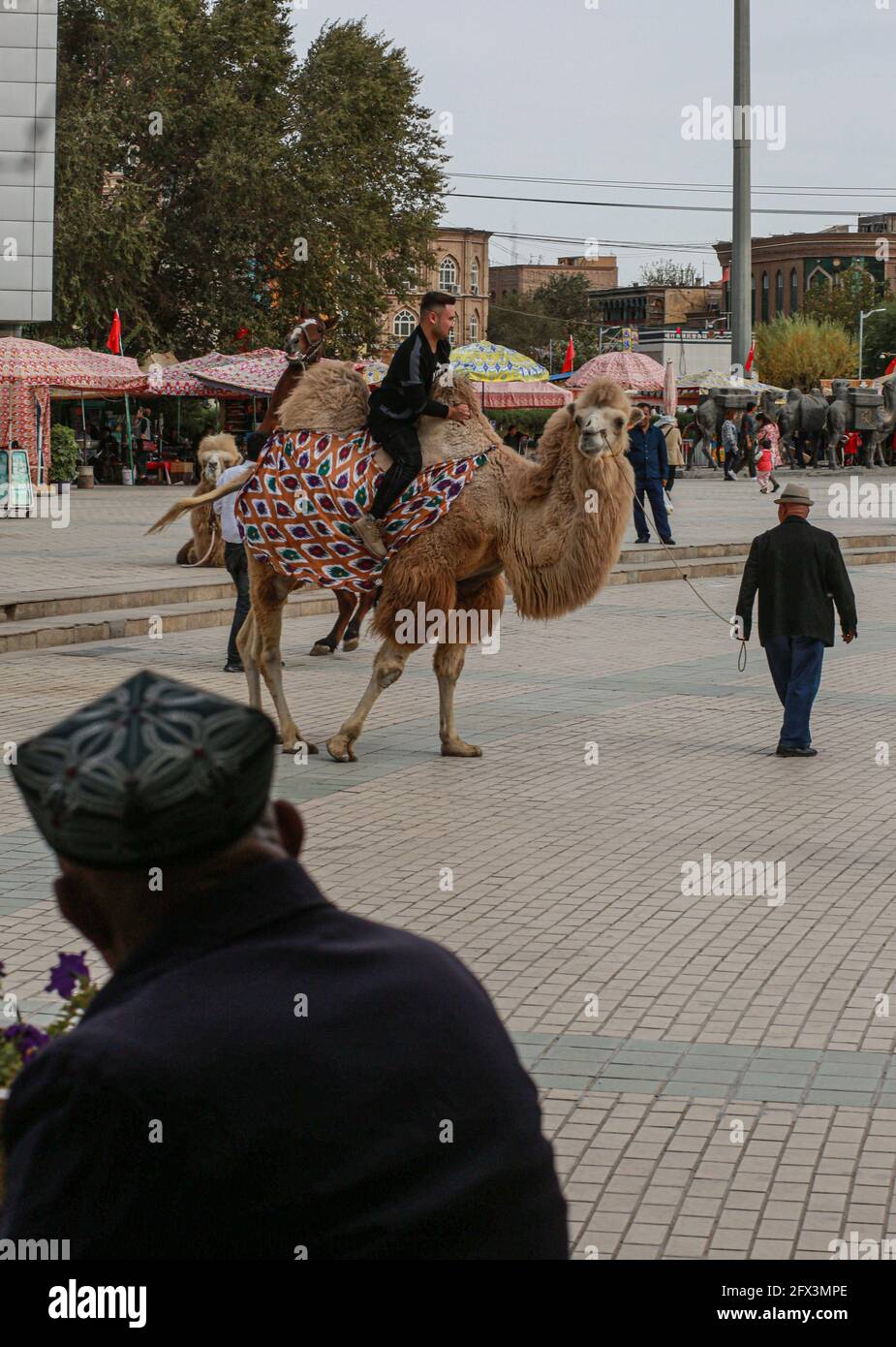 Vecchio uomo visto dalla parte posteriore con il cappello tradizionale di Uihgur guardando un turista che cavalcano un cammello. Kashgar, Repubblica popolare cinese Foto Stock
