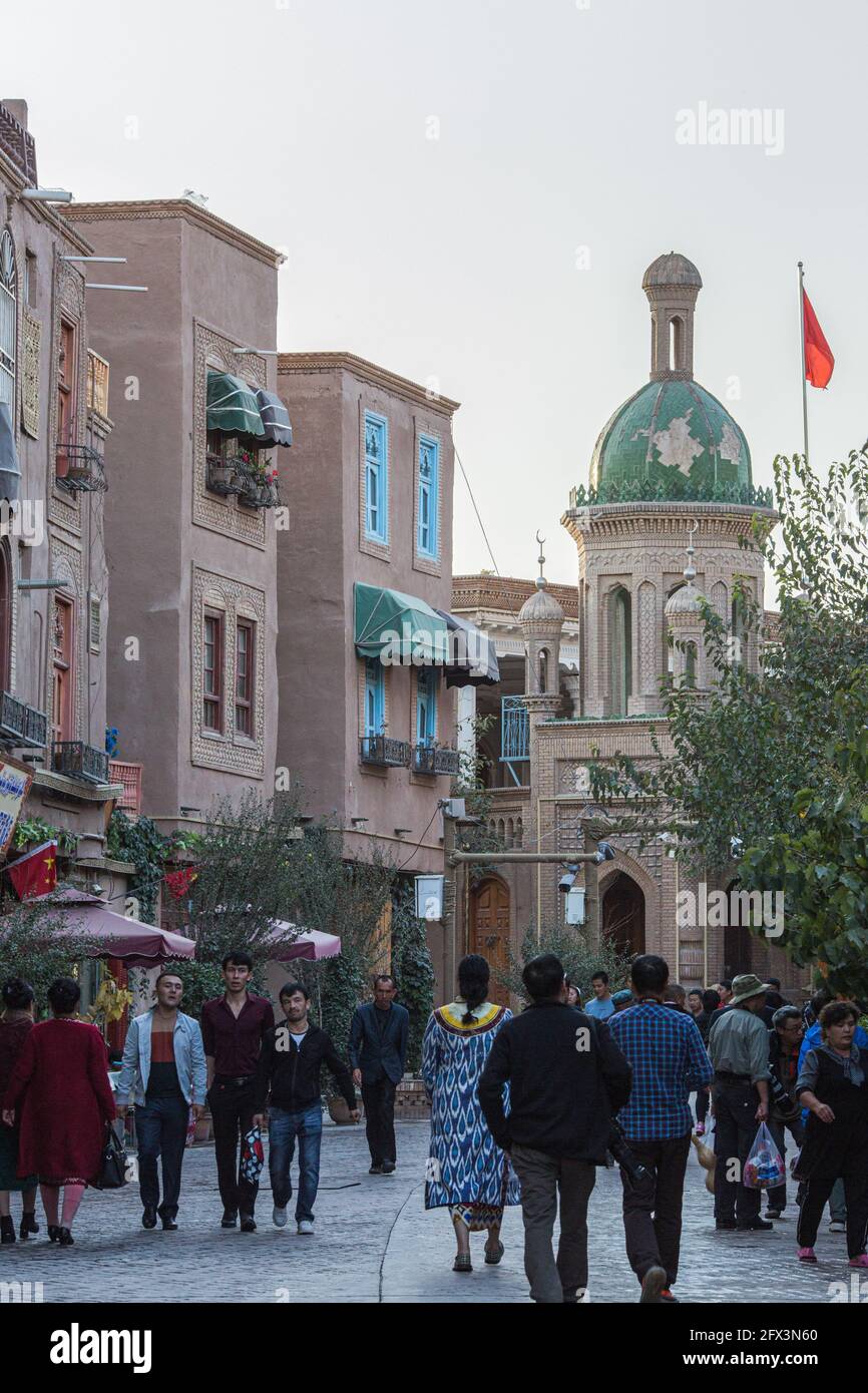 La gente cammina lungo una strada a Kashgar, sullo sfondo vediamo un tempio islamico e la bandiera della Repubblica popolare Cinese sventolare Foto Stock