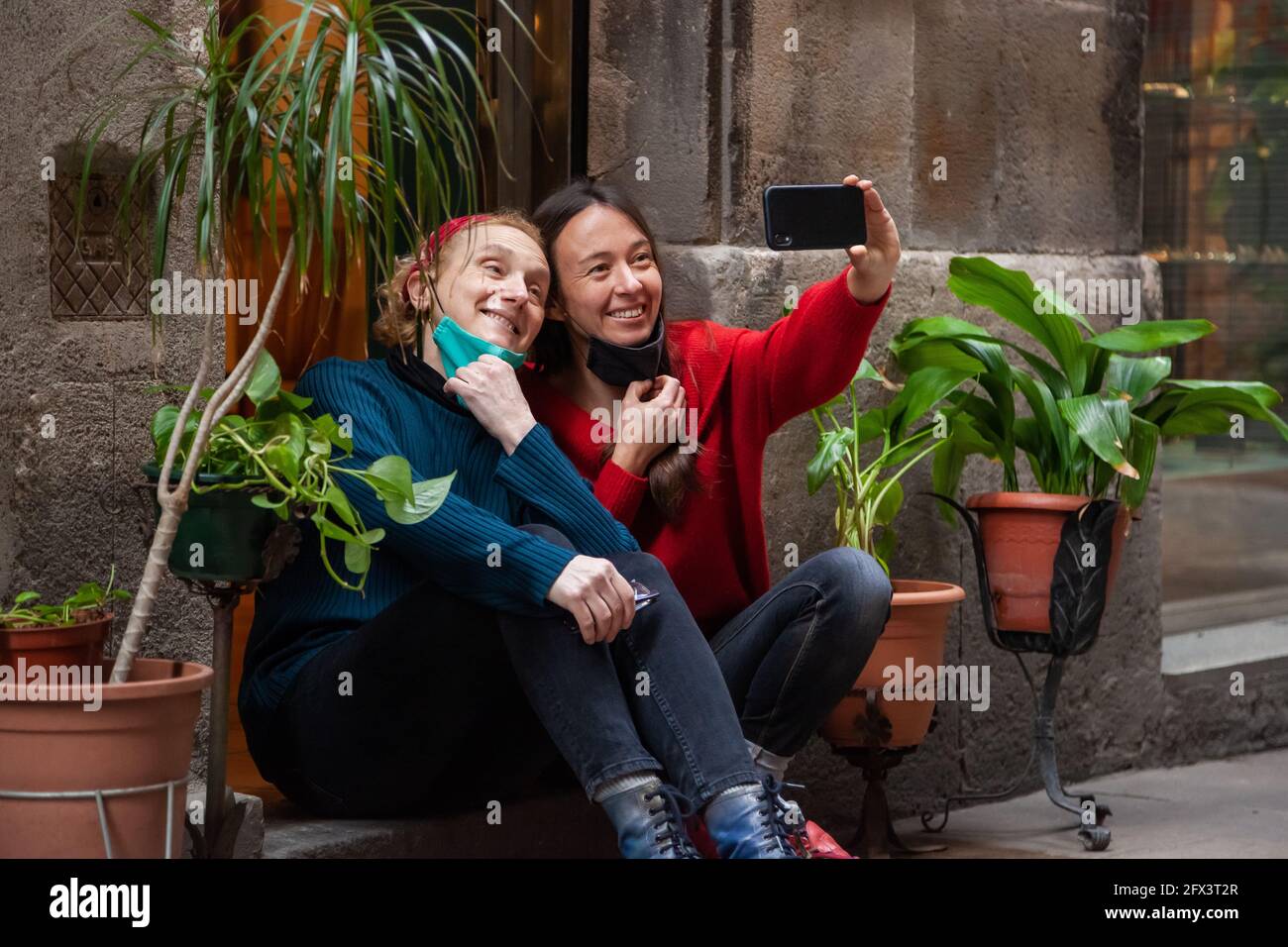 Due amici di donna che indossano maschere di protezione seduta vicino insieme prendendo selfie con smartphone. Concetto: Stile di vita cittadino, covid-19, tutti i giorni noi Foto Stock